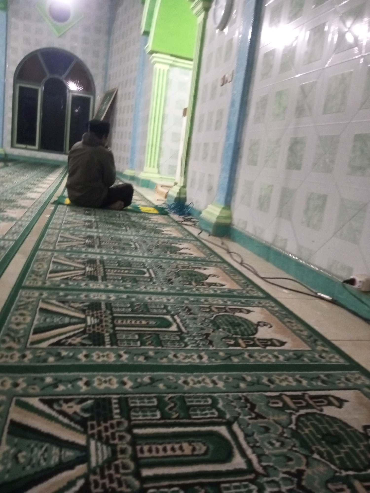 The image shows a man wearing a dark jacket and black pants sitting on a prayer rug in what appears to be a mosque or prayer room. The man is facing away from the camera. The prayer rug has a green and beige geometric pattern. The walls are tiled with a light green and white pattern, and there are blue and green architectural elements visible. A large arched window with dark panes is visible on the left side of the image. The lighting suggests it is indoors, likely evening or early morning, as the light sources are artificial and diffuse. The scene appears to be located in a public or semi-public building, consistent with a place of worship. No specific details about the city of Andongsili, Indonesia, are discernable from the image itself.