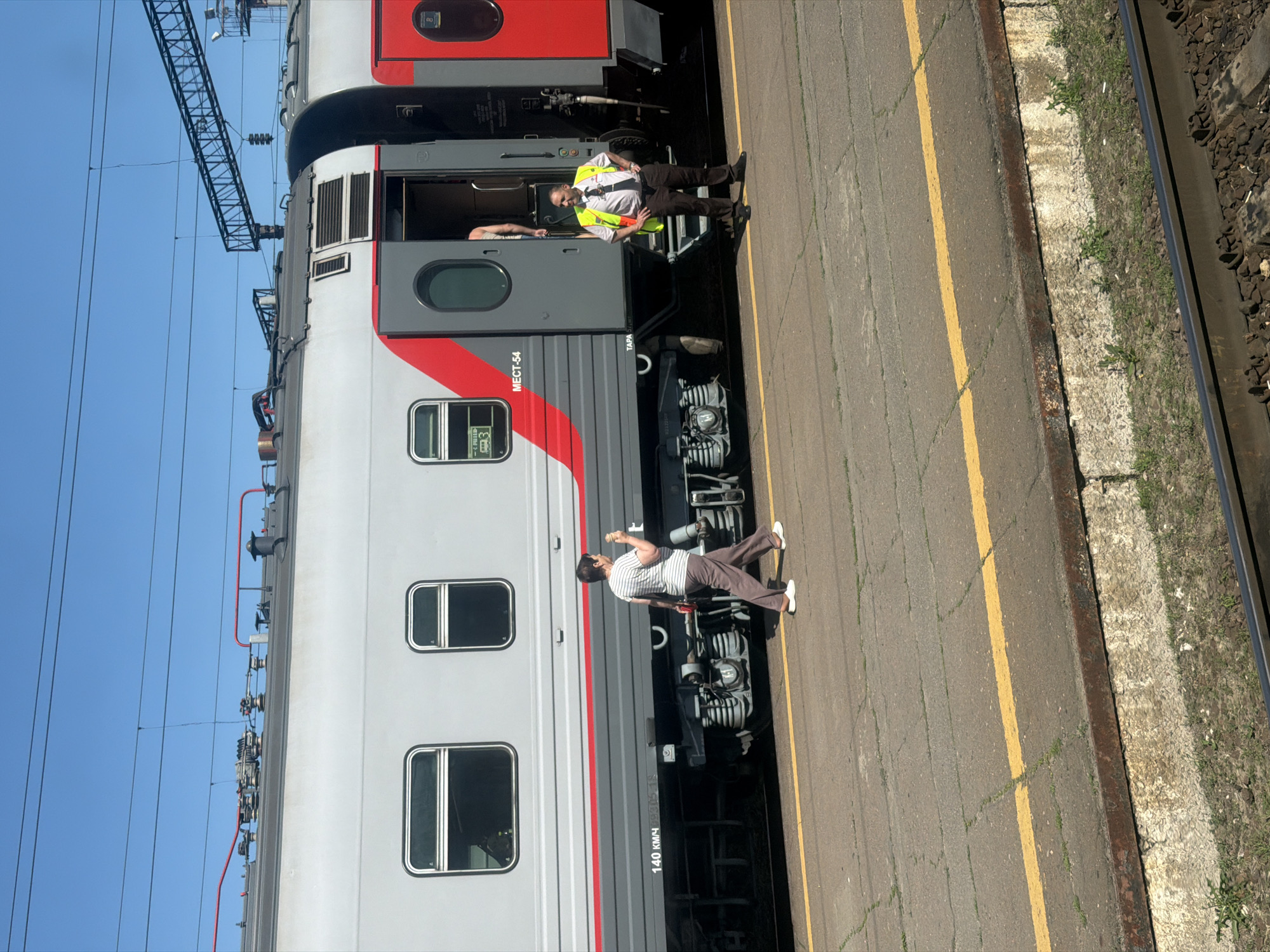 In Лиски, Russia, a train car with gray and red accents sits at a platform. A conductor in a yellow vest and dark pants stands by the open train door. A woman in a striped shirt and brown pants walks away from the train, holding an ice cream cone. The platform is made of concrete slabs with yellow lines marking the edge. The sky is clear and blue.