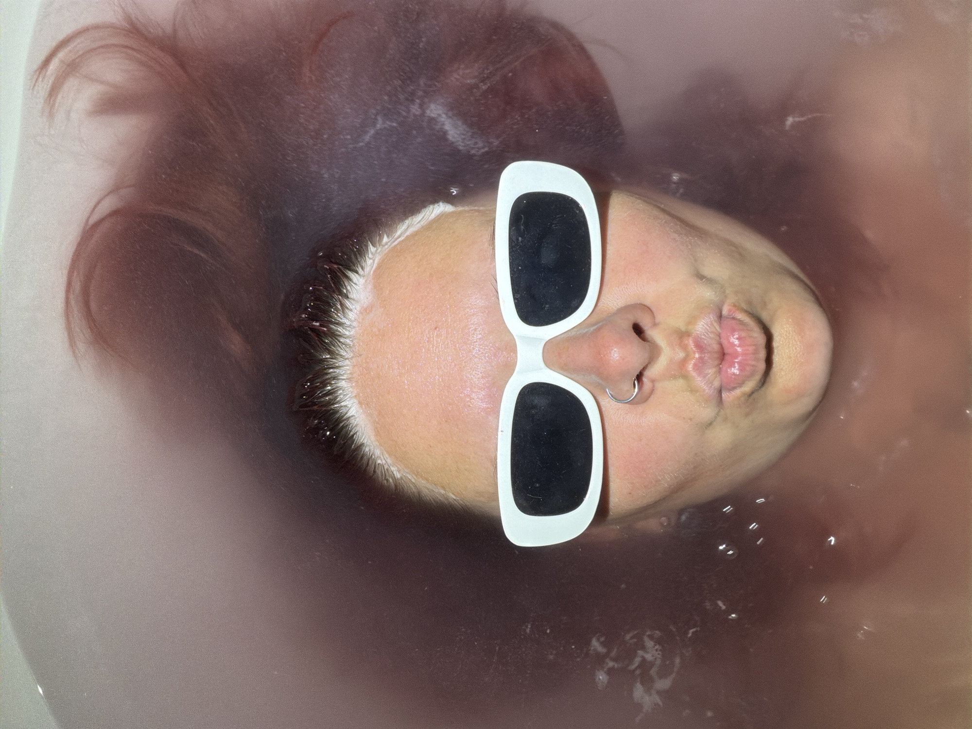 A person with auburn hair is submerged in a tub of murky water. They are wearing oversized white sunglasses with black lenses. A silver hoop earring is visible in their left nostril. The overall environment appears to be a bathroom.