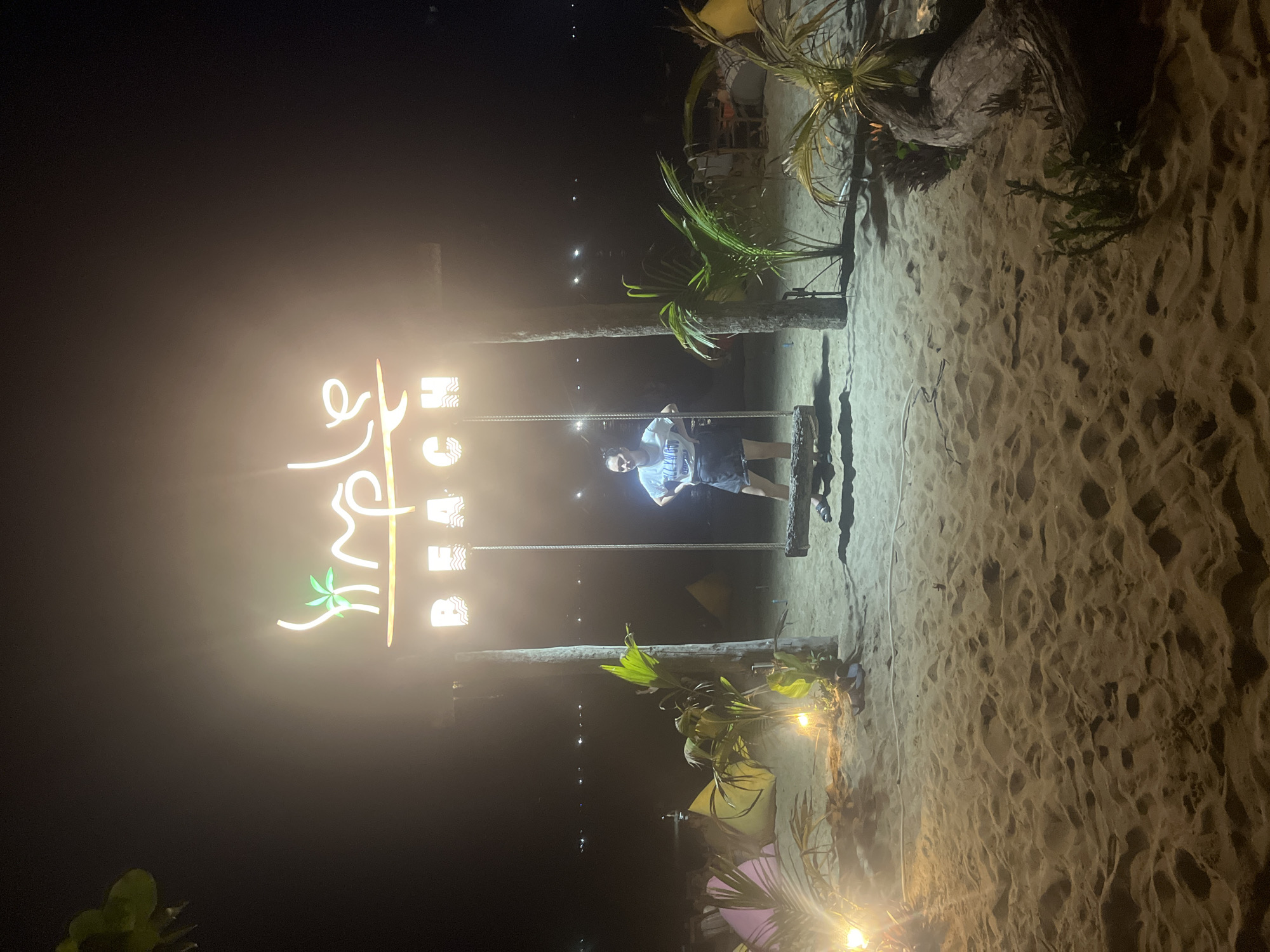 The image depicts a beach at night. A woman stands on a swing, which is suspended between two wooden posts. Above the swing, a neon sign reads "Simple Beach" with a palm tree illustration. The beach is sandy, with scattered palm trees and some tents in the background. Lights illuminate the scene, casting a warm glow on the sand and foliage. The ocean is visible in the distance, with faint lights reflecting on its surface. The overall atmosphere is tropical and relaxed. This appears to be a scene from a beach resort or a public beach with amenities in Da Nang, Vietnam.