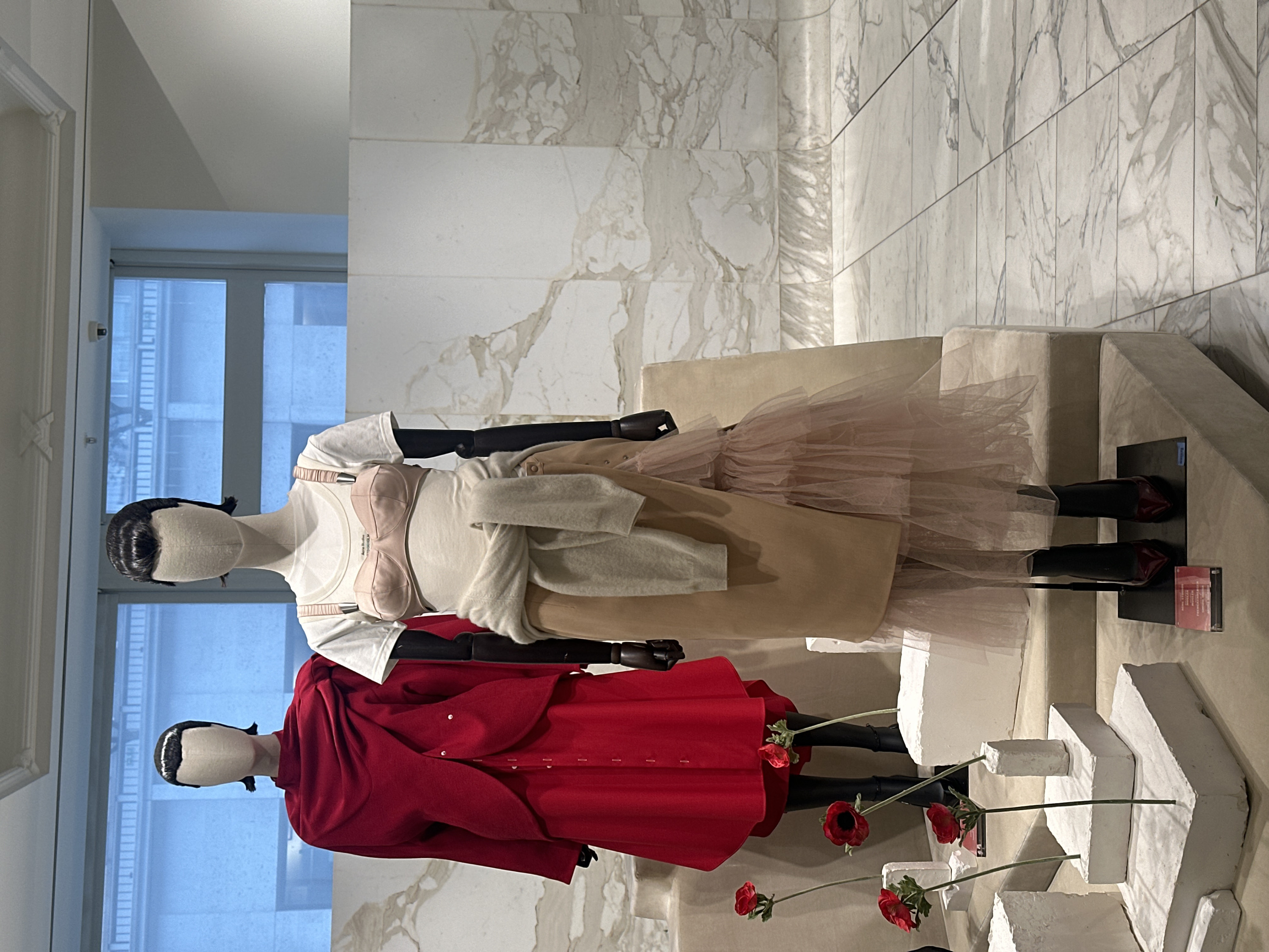 This image depicts a static fashion display inside a retail establishment, likely a high-end boutique or department store, located in the city of London, United Kingdom. The scene features two headless, faceless mannequins presented on multi-tiered, fabric-covered platforms against a backdrop of a large window and marble-clad walls.
The mannequin on the left, positioned closer to the viewer, wears a striking red ensemble consisting of a long-sleeved top or jacket with a high collar and a draped, asymmetric front closure secured by two white buttons, over a matching red A-line skirt or dress with a line of small white buttons down its front. This mannequin's legs are clad in black boots.
The mannequin on the right, standing slightly further back, is dressed in a layered outfit. It wears a white short-sleeved t-shirt with "Acne Studios Stockholm" printed on the chest, overlaid with a light pink satin bra featuring wide shoulder straps with metallic buckles. A light beige, possibly wool or felt, asymmetric wrap skirt is worn over a pale pink, ruffled tulle skirt that extends beneath it. A light beige or cream-colored sweater is tied around this mannequin's waist. Its feet are adorned with dark red, pointed-toe patent heels, resting on a black rectangular base with a transparent information plaque displaying text that includes "MUGLE", "MULLER", "FRENCHENBERGER", and "ACNE STUDIOS".
Several white, irregularly shaped block-like structures are arranged on the display platforms, around the mannequins' feet. Red artificial anemone flowers with green stems are interspersed among these blocks, with some stems inserted into smaller, white vase-like blocks.
The environment behind the mannequins features a large rectangular window revealing a bright blue sky and the light-colored stone facade of a classical building with visible windows and architectural elements, characteristic of central London architecture. To the right of the window, the interior wall is covered in white marble tiles with prominent grey and brown veining, which continues onto the tiled floor. The ceiling is white with decorative molding, and a small black security camera is visible in the upper right quadrant of the image. The overall lighting is bright and even.