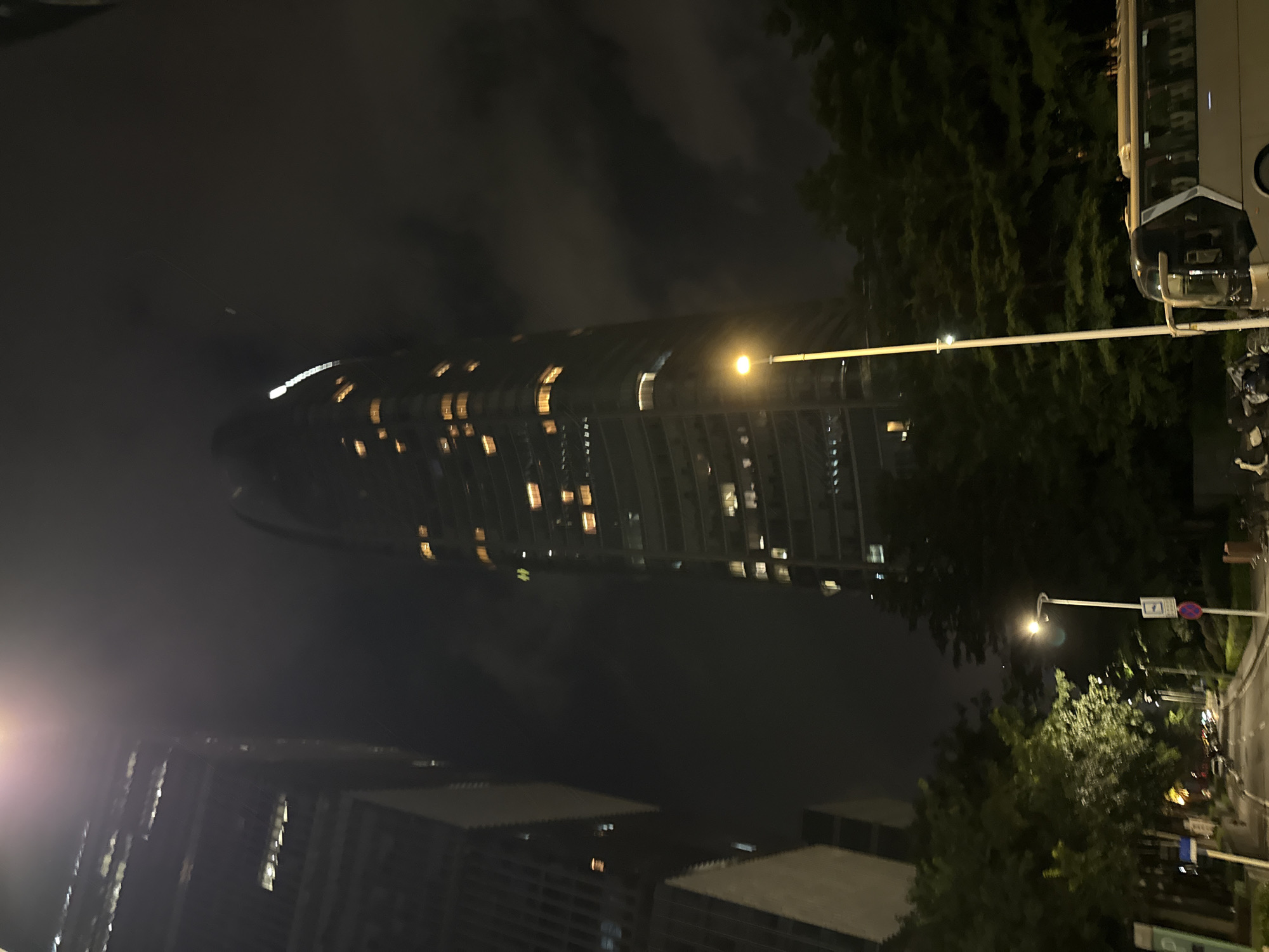 The image depicts a nighttime scene in Haizhu District, China, featuring a prominent, illuminated skyscraper with a curved facade. The building's windows glow with warm light, contrasting with the dark, cloudy sky. In the foreground, trees and streetlights cast a soft glow on a street that appears to be in a relatively quiet urban area. The presence of a bus on the right side of the image suggests a transportation hub or a road frequented by public transport.
The overall atmosphere is one of urban tranquility at night, with the city lights creating a sense of depth and scale. The low-hanging clouds add a touch of drama to the scene, partially obscuring the top of the skyscraper and creating a moody ambiance. There are no visible people in the frame, and the focus is primarily on the architectural elements and the urban landscape.
Notable details include the time of day, clearly night, and the presence of clouds which contribute to the overall mood. The architecture of the skyscraper, with its unique shape and illuminated windows, is a key focal point. Streetlights and trees further define the setting, indicating an outdoor urban environment. The image appears to be taken from a slightly elevated perspective, looking up at the skyscraper. No discernible text is visible in the image.