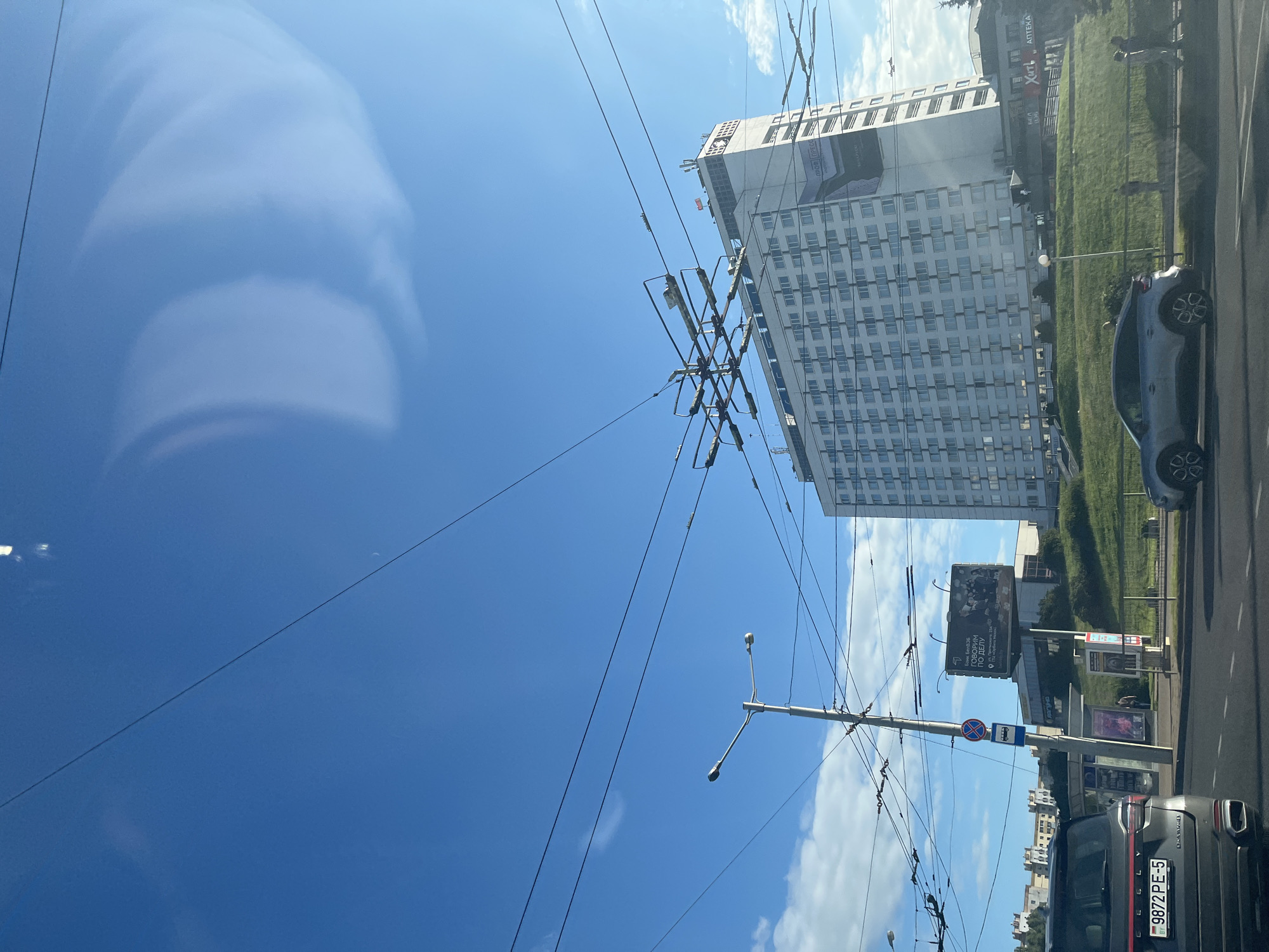 The image shows a street scene in Minsk, Belarus, on a clear day with a bright blue sky and scattered clouds. Dominating the right side of the frame is a large, white, multi-story hotel building with many windows. In front of the hotel, a grassy incline is visible. Overhead, a complex network of tram or trolleybus wires crisscrosses the sky.
On the left side of the image, a black SUV with a Belarusian license plate reading "9872 PE-5" is parked on the road. Next to it, a silver hatchback is also on the road. A street light pole with multiple fixtures stands prominently in the mid-ground. A bus stop sign is visible on the pole, along with a large billboard displaying text and images. Further in the background, other buildings and trees can be seen. Several individuals are present, walking on the sidewalk or near the bus stop. The overall impression is a typical urban environment with active street life.