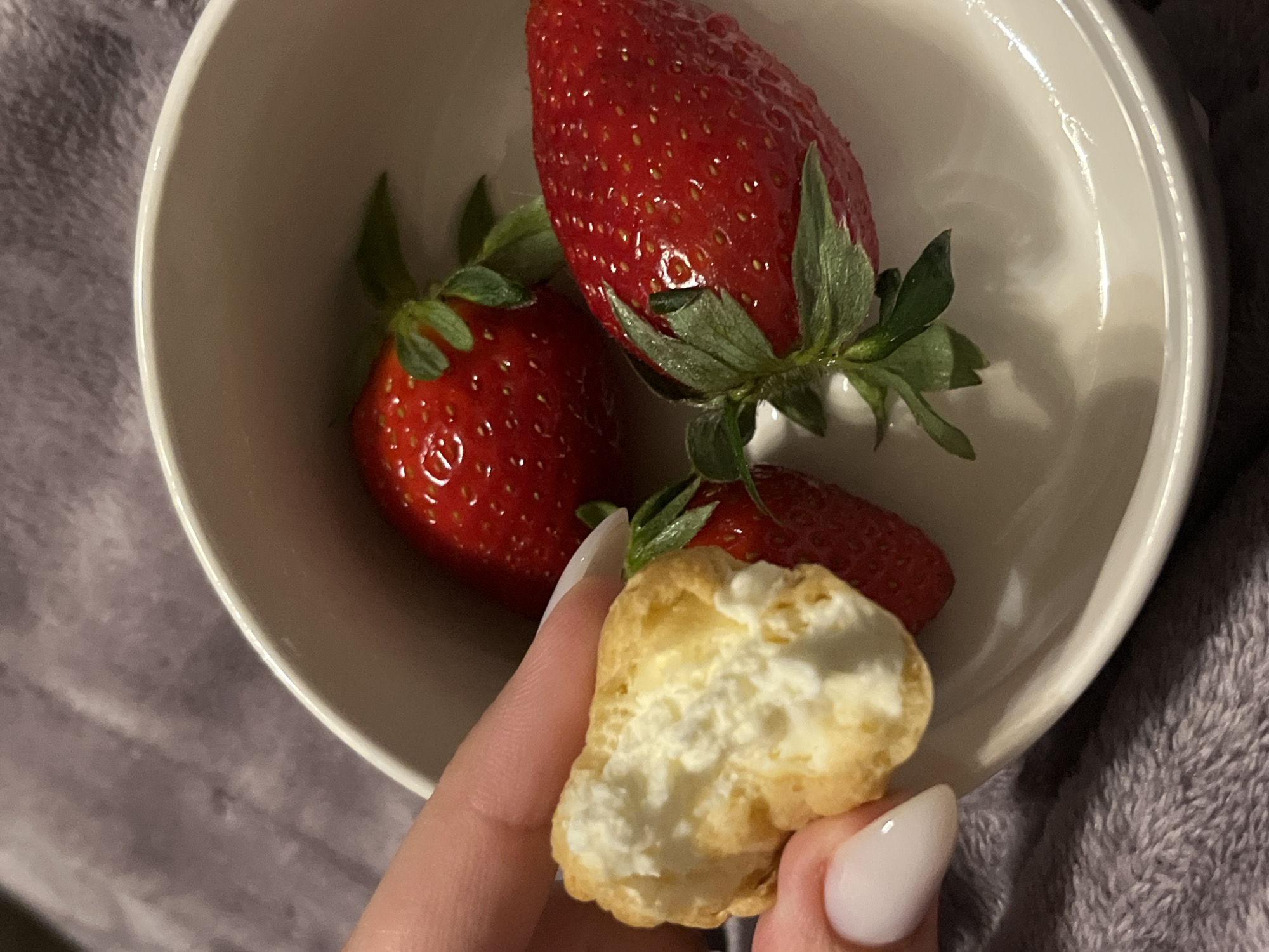 A top-down, indoor shot shows a white bowl containing three strawberries with green stems. A partially eaten cream puff is held in a hand with manicured fingernails, positioned in front of the bowl. The bowl rests on a grey, textured surface. The location is unknown and not specified as Sofia, Bulgaria.