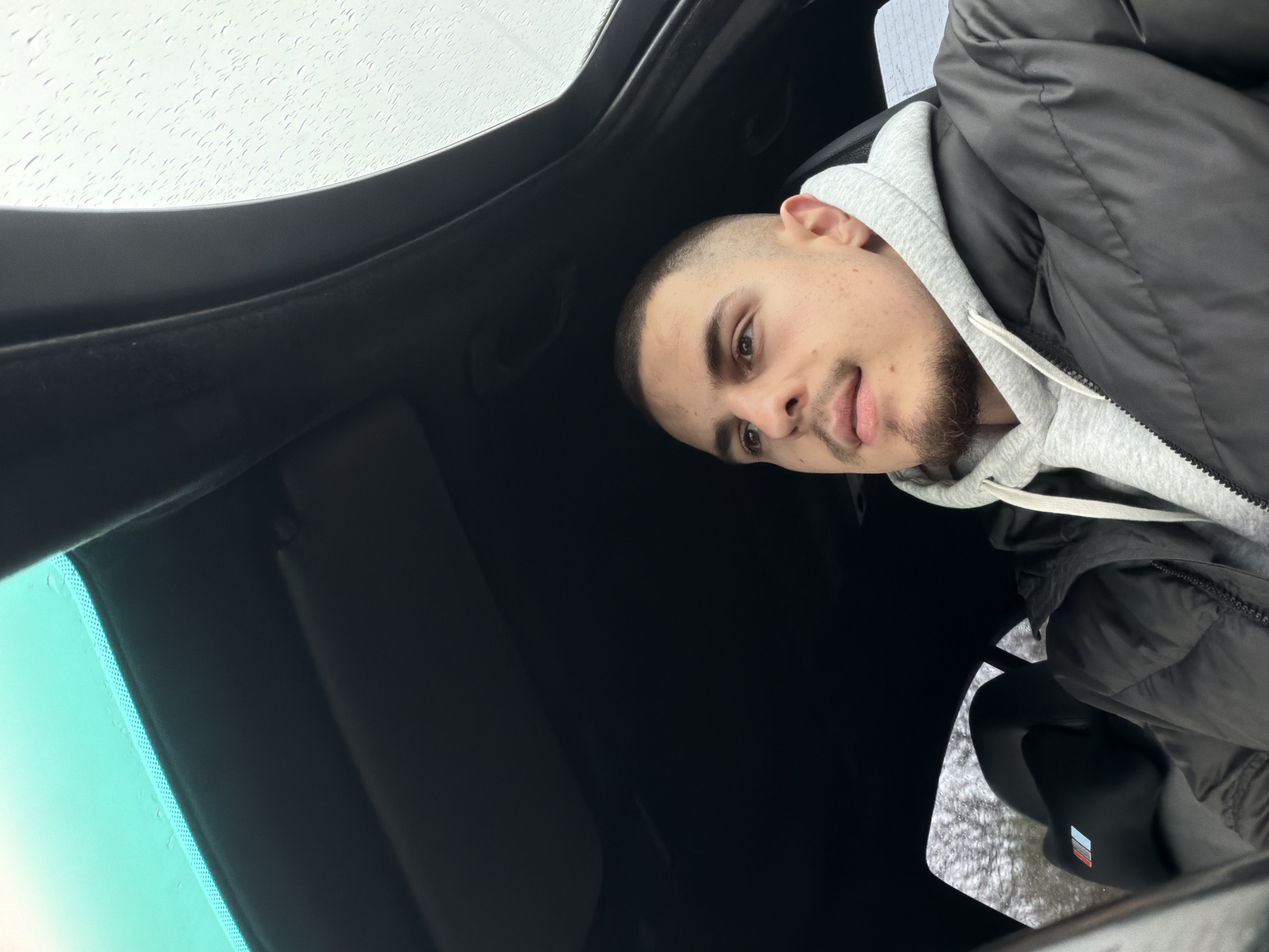 A man with short brown hair and a light beard is sitting in a car, looking towards the camera with his tongue slightly protruding. He is wearing a grey hooded sweatshirt and a black puffy jacket. The car has a sunroof, visible in the background with raindrops on the glass. The interior of the car is black. The location is likely Plovdiv, Bulgaria, but this cannot be confirmed from the image alone. The focus is on the man and the interior of the vehicle.