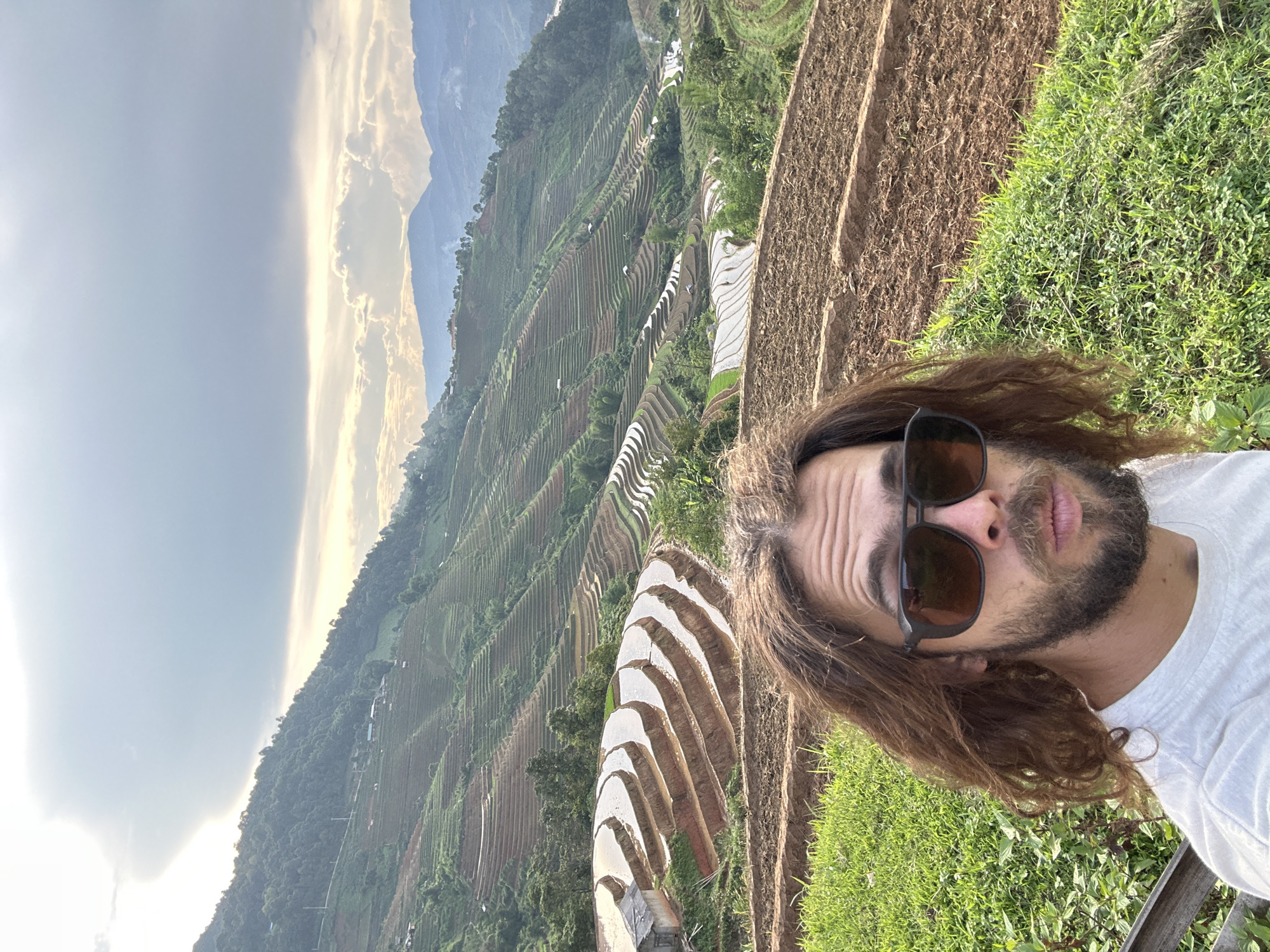 A man with long, wavy brown hair, a beard, and dark sunglasses takes a selfie in the foreground, his face angled slightly towards the camera with a neutral expression. He is wearing a white t-shirt. The immediate foreground also shows green grass and patches of brown earth.
Behind him, a breathtaking landscape of terraced rice paddies unfolds across a mountainous region, characteristic of Lao Chai, Vietnam. The terraces are carved into the hillsides, with many segments appearing to be filled with water, reflecting the soft light from the sky. Other sections show cultivated land or sparse green vegetation. In the distance, layers of mountains rise under a dramatic, partly cloudy sky. The lighting, with warm hues reflecting off the clouds and illuminating the scene, suggests either sunrise or sunset. There is no visible text in the image.