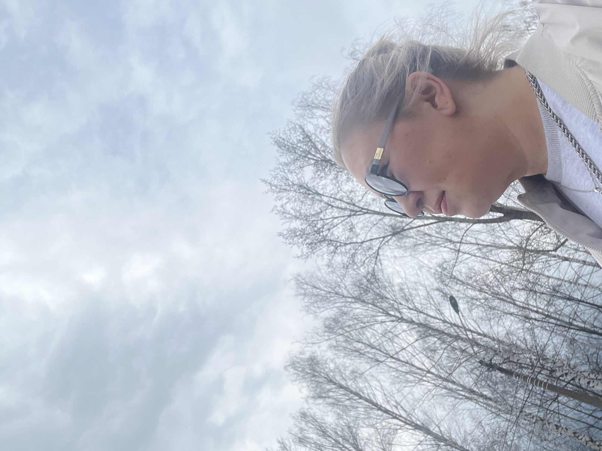 The image shows a person with blonde hair, wearing sunglasses, a light-colored jacket, and a silver chain. The person is looking down. The background includes bare tree branches against a cloudy sky. The location is potentially in Chaikovsky, Russia. There is also a streetlight in the background.