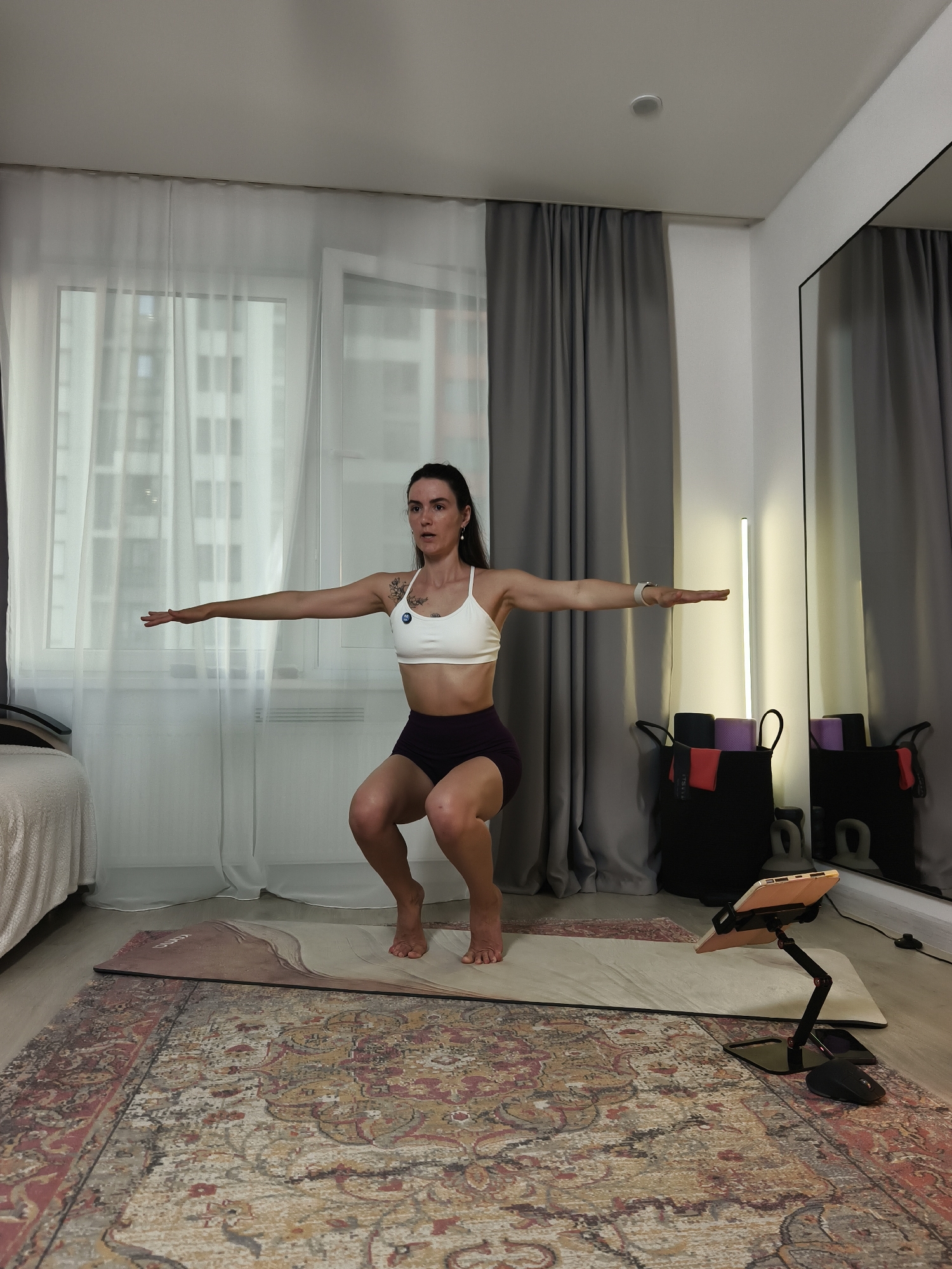 A woman is performing a squat exercise in a well-lit room, likely an apartment in the city of Tyumen, Russia. She is wearing a white sports bra and dark shorts, with her arms extended outwards for balance. The room features large windows with sheer white curtains, gray floor-to-ceiling drapes, and a large floor rug with an intricate pattern. In the background, a full-length mirror reflects the scene, and a tall, thin lamp stands near a basket filled with yoga mats and other exercise equipment. A yoga mat is laid out on the floor in front of the woman, and a tablet on a stand is positioned nearby, suggesting she might be following an online workout. A bed with a textured white cover is visible on the left side of the image.