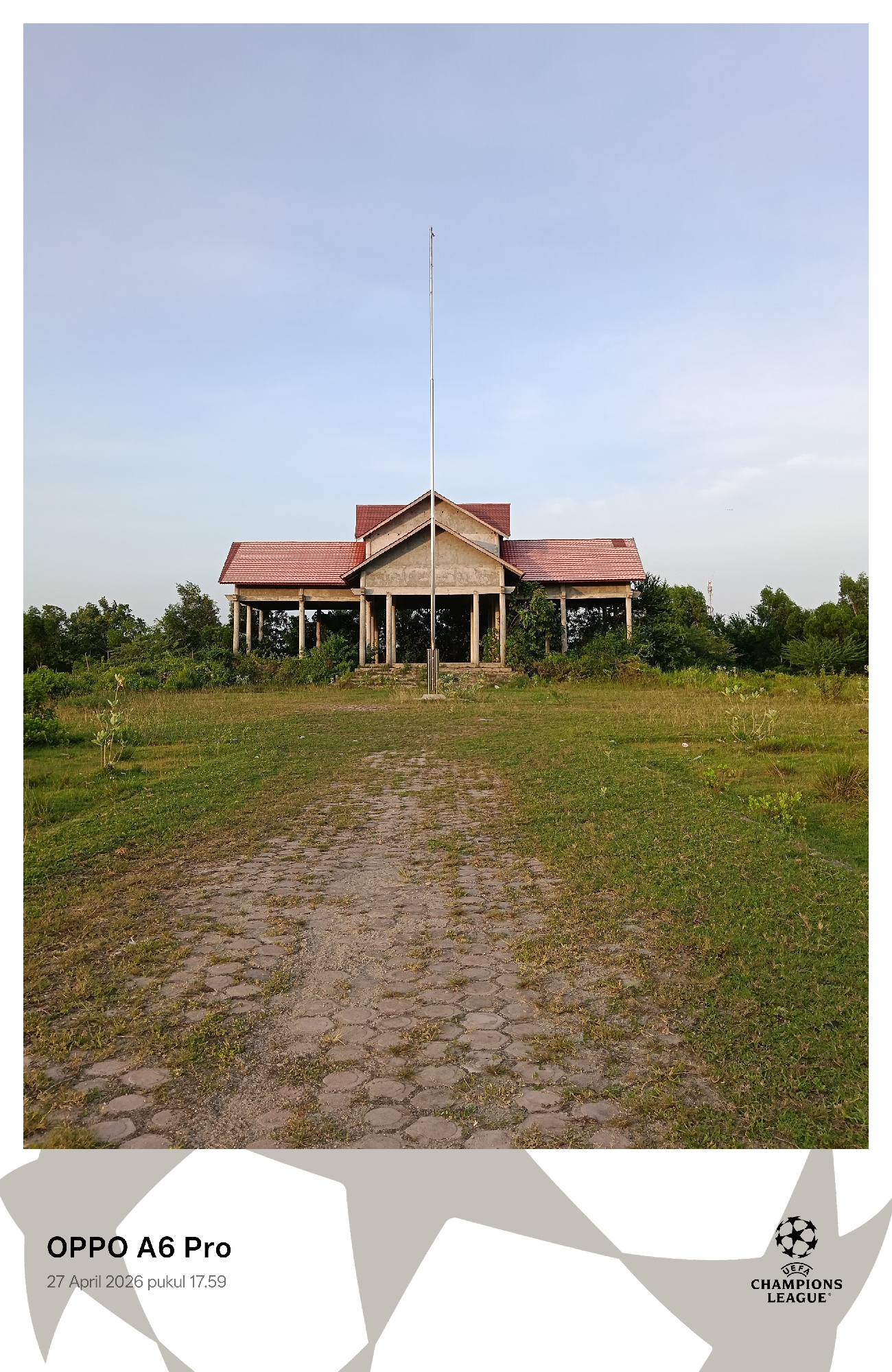 The image depicts an unfinished or abandoned structure located in Lhokseumawe, Indonesia. The building has a red-tiled roof and concrete pillars, with greenery growing around it. A tall, metal pole stands directly in front of the building. A path of hexagonal paving stones leads from the viewer to the structure. The surrounding area is overgrown with grass and small shrubs. The sky is clear and pale blue. The bottom of the image includes the text "OPPO A6 Pro" and "27 April 2026 pukul 17.59," along with the UEFA Champions League logo.