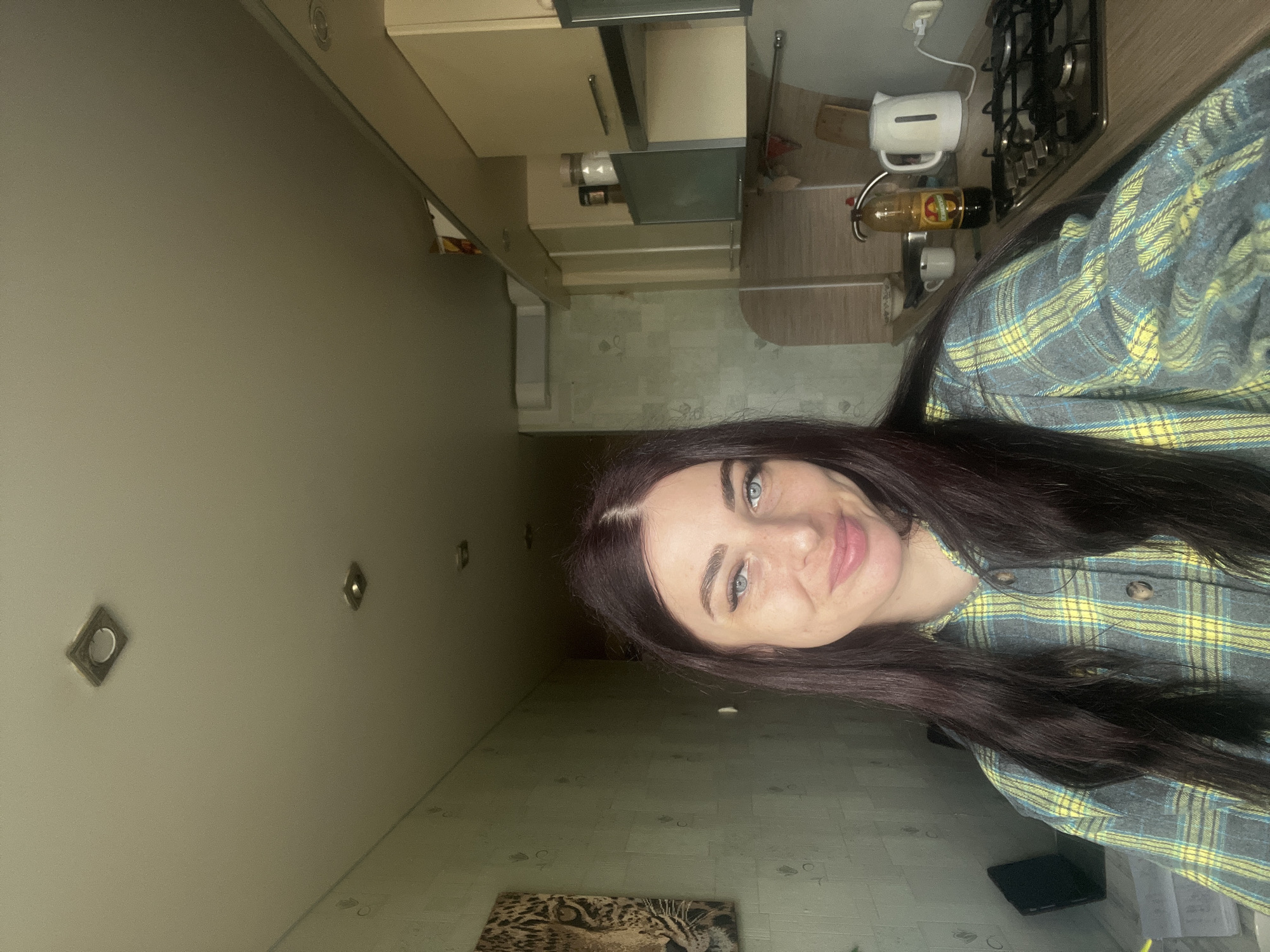 A selfie-style image captures a woman with long, dark, slightly wavy hair and striking blue eyes, looking towards the camera with a gentle, pleasant smile. She is wearing a plaid shirt patterned with yellow, light blue, and dark blue/gray colors. The image is presented sideways, suggesting it was taken as a vertical selfie.
The background depicts a domestic kitchen environment, consistent with a home in Chaikovsky, Russia. On the right side of the frame, behind the woman's shoulder, a kitchen countertop is visible. It holds a white electric kettle, a dark gas stove with black grates, and an amber-colored bottle with indistinct text that appears to be a brand name for a liquid like oil or syrup, along with two white mugs. Above the counter are light-colored kitchen cabinets and an extractor hood. The walls are a light, neutral shade of gray or beige, and several square recessed spotlights illuminate the ceiling. The floor, visible in the lower portion of the image, has a light-colored, possibly patterned or textured surface. In the bottom left corner of the frame, a picture or canvas featuring a detailed close-up of a leopard's face is resting, possibly on the floor or a low surface. The lighting is artificial, suggesting an indoor setting without strong natural light cues for the time of day.