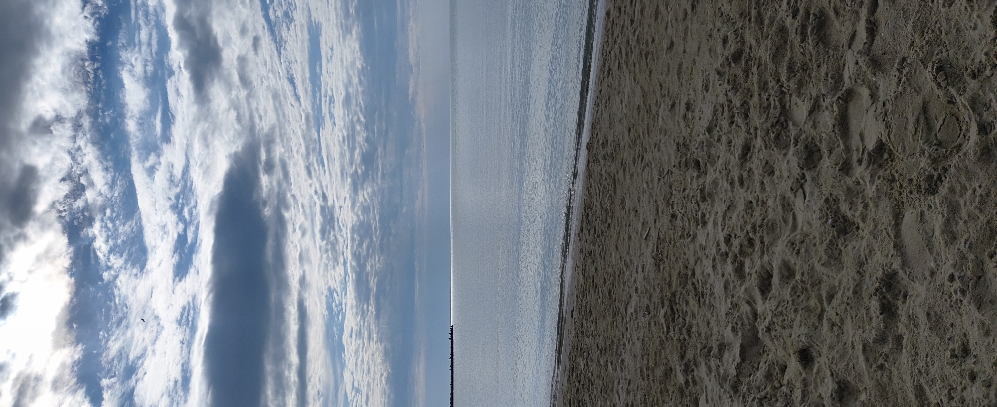 The image shows a beach scene. On the left is a sky filled with clouds, ranging from white to dark gray, with a glimpse of the sun. In the middle is the calm sea, with a slight glare and a distant breakwater structure. On the right is a sandy beach with numerous footprints. The image likely captures a scene on the beach of Varna, Bulgaria.