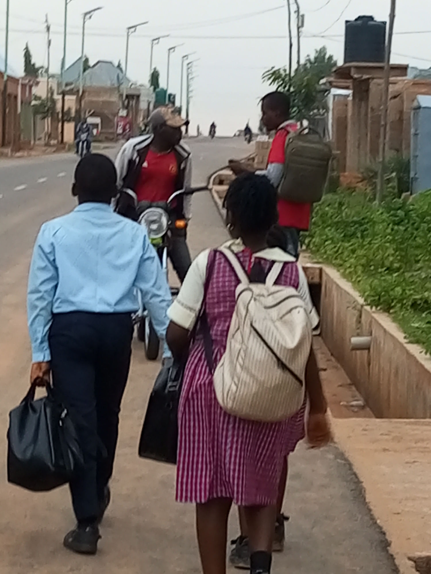 The image depicts a street scene in Gombe, Nigeria. In the foreground, three individuals are walking away from the camera on a paved pathway. The person on the left is a male wearing a light blue long-sleeved shirt and dark pants, carrying a black duffel bag in his left hand. Next to him, a young female is walking, wearing a striped pink and white dress and a light-colored backpack with vertical stripes. To the right of the female, partially visible, is another person. Behind the foreground individuals, a man in a red shirt and a camouflage baseball cap is seated on a motorcycle, facing towards the right. The motorcycle is facing the camera and appears to be in motion or recently stopped. Further in the background, other people on motorcycles and a few buildings are visible. The environment appears to be an urban or semi-urban setting with streetlights lining the road. The overall lighting suggests it is daytime, possibly overcast.