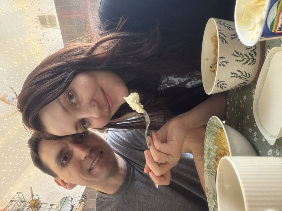 A smiling couple poses close together. The woman holds a fork with a dollop of white food, possibly cream or frosting. Bowls of cereal and a container of something creamy are visible on the table. The background features a lit string of lights and a wall with subtle patterned wallpaper. The overall scene suggests a casual, at-home setting, potentially during a meal or snack time.