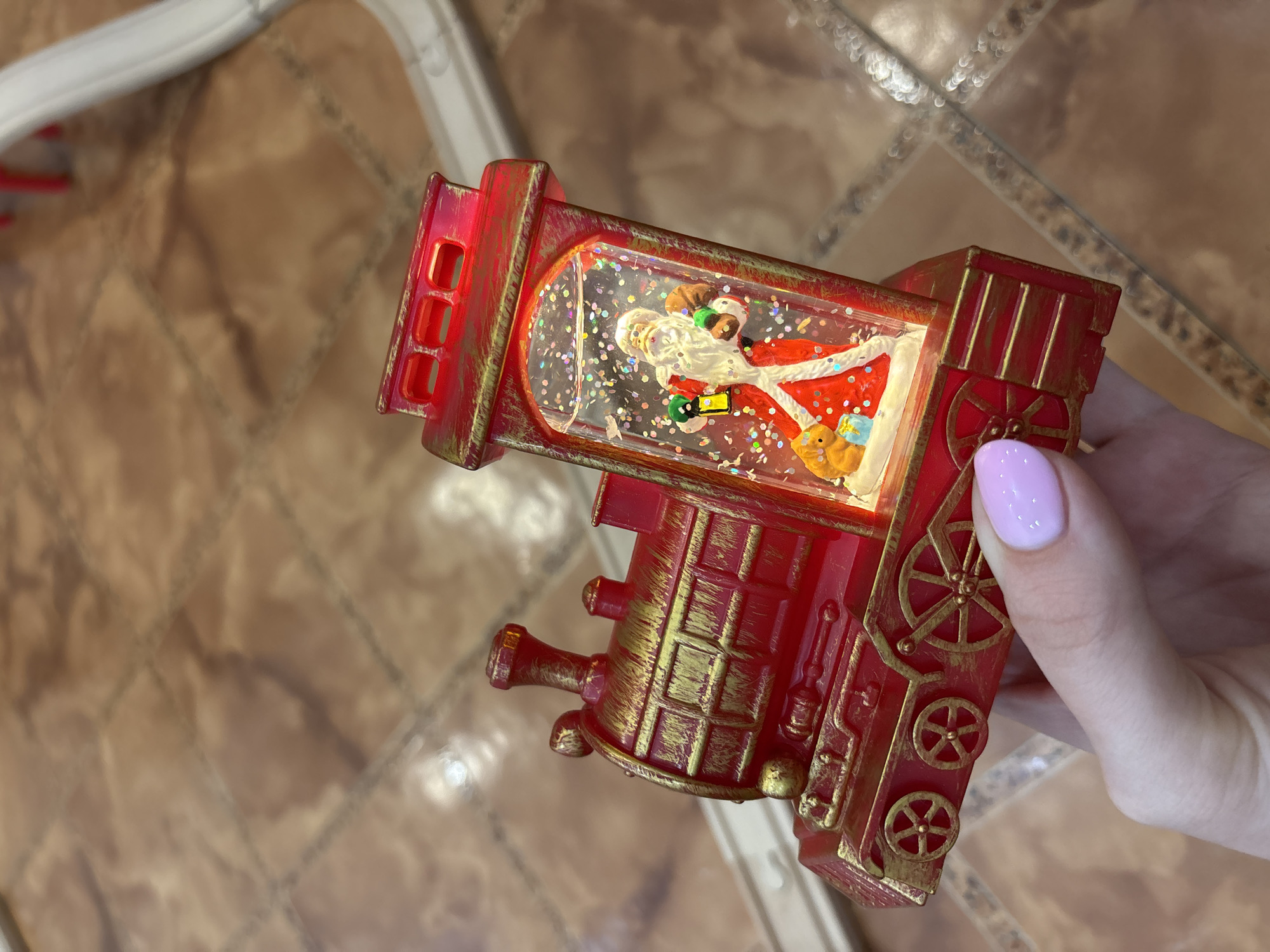 A hand holds a red and gold toy train with a snow globe containing a Santa Claus figure. The background shows brown and tan patterned floor tiles.