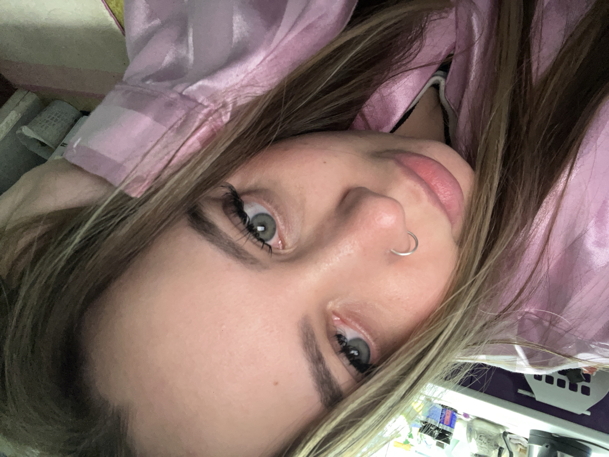 A close-up selfie of a fair-skinned woman with light brown hair, wearing a pink, satin-like top. She has a silver hoop nose piercing and is looking directly at the camera. The background appears to be an indoor setting with various objects and containers visible, although indistinct, suggesting a somewhat cluttered environment. There is no information to suggest this photo was taken in Novopokrovskaya, Russia.