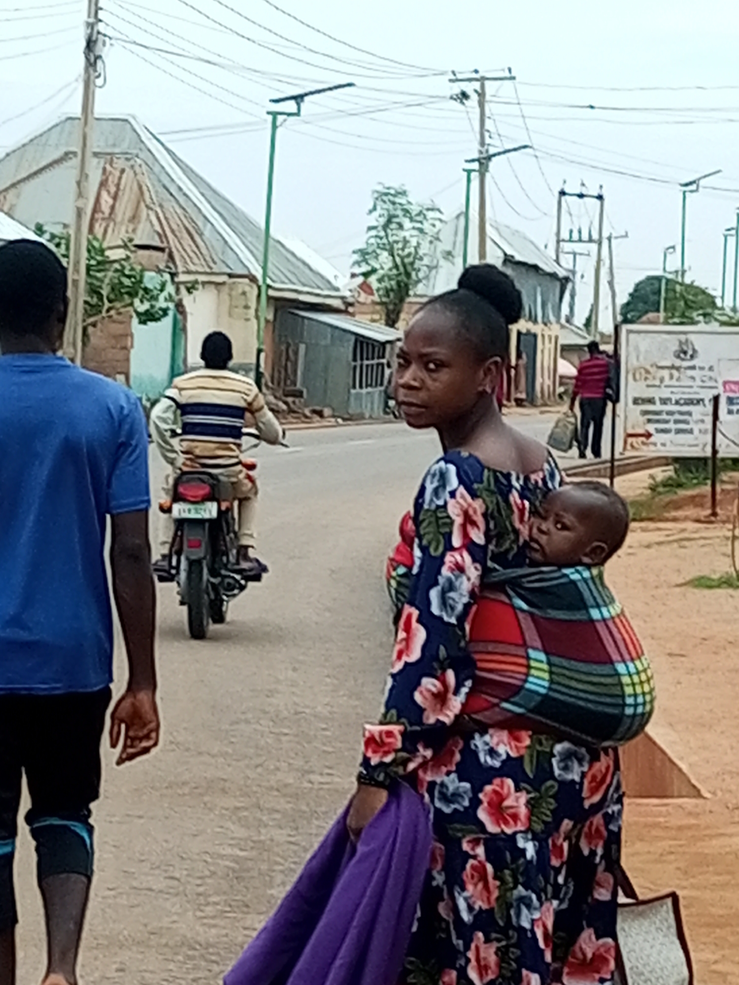 A medium shot captures a street scene in Tempure, Nigeria, under an overcast sky. In the foreground, a woman with dark skin and her hair in a bun faces left, looking at the camera. She wears a navy blue dress with a floral pattern featuring red and pink flowers and green leaves. A baby is strapped to her back in a colorful, patterned wrap. To the left, a person seen from behind wears a blue t-shirt and black shorts with a blue stripe. Further down the road, a person on a motorcycle travels away from the camera, wearing a striped shirt and shorts. In the background, buildings with corrugated metal roofs and some trees are visible. A street sign with text and an emblem stands on the right side of the frame. The ground is paved, appearing to be a road.