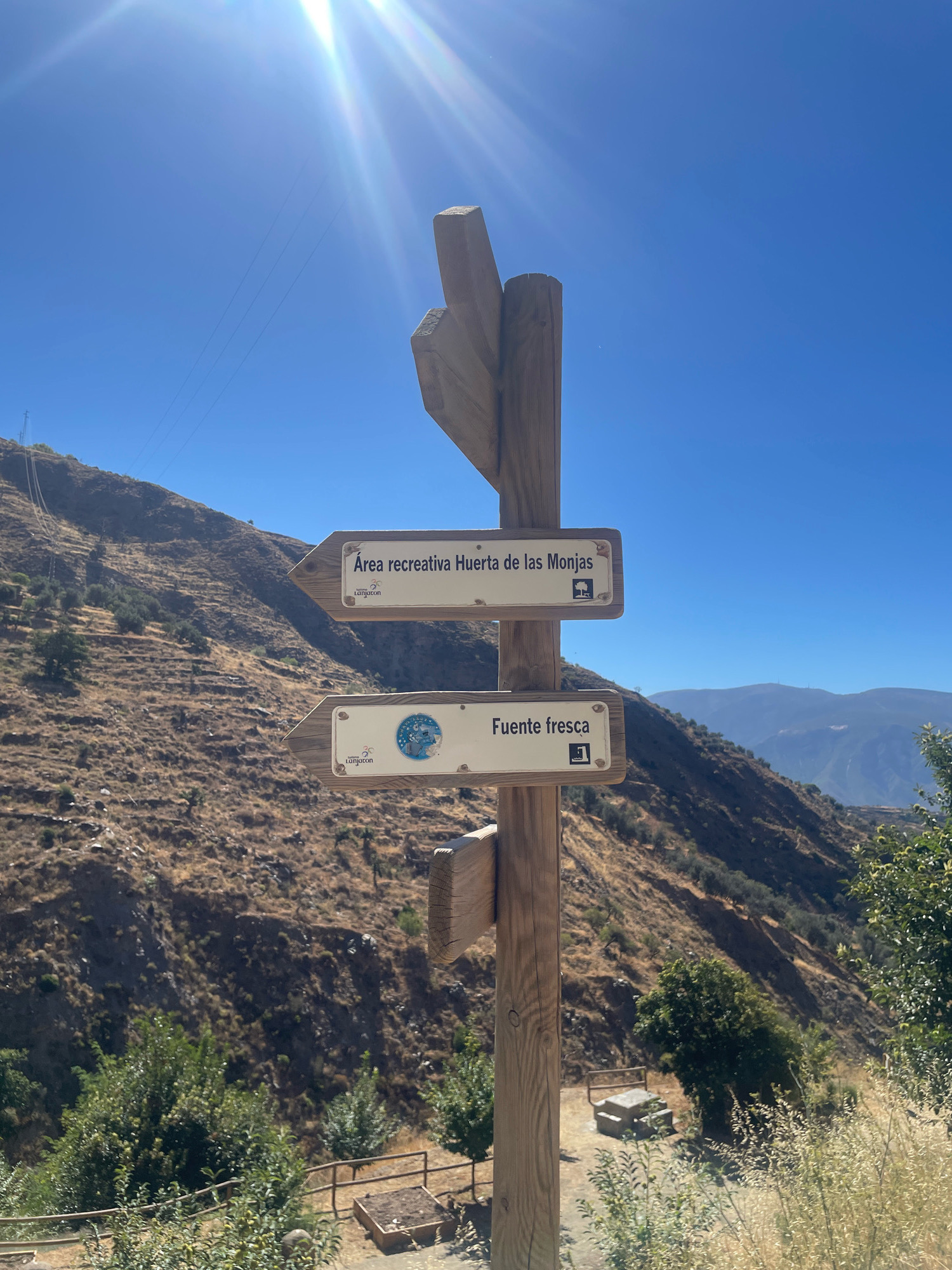 A wooden signpost stands against a bright blue sky, its directional signs pointing towards a recreational area and a fresh spring. The signpost is positioned on a hillside dotted with dry vegetation and scattered trees. In the background, distant mountains are visible. The sign has two planks, each with a sign attached. The top sign reads "Área recreativa Huerta de las Monjas" and has an arrow pointing left. The bottom sign reads "Fuente fresca" and has an arrow pointing right. Both signs feature a logo with the text "Lanjarón". There are no people visible in the image. The environment suggests a natural, possibly mountainous area in or near Lanjarón, Spain.