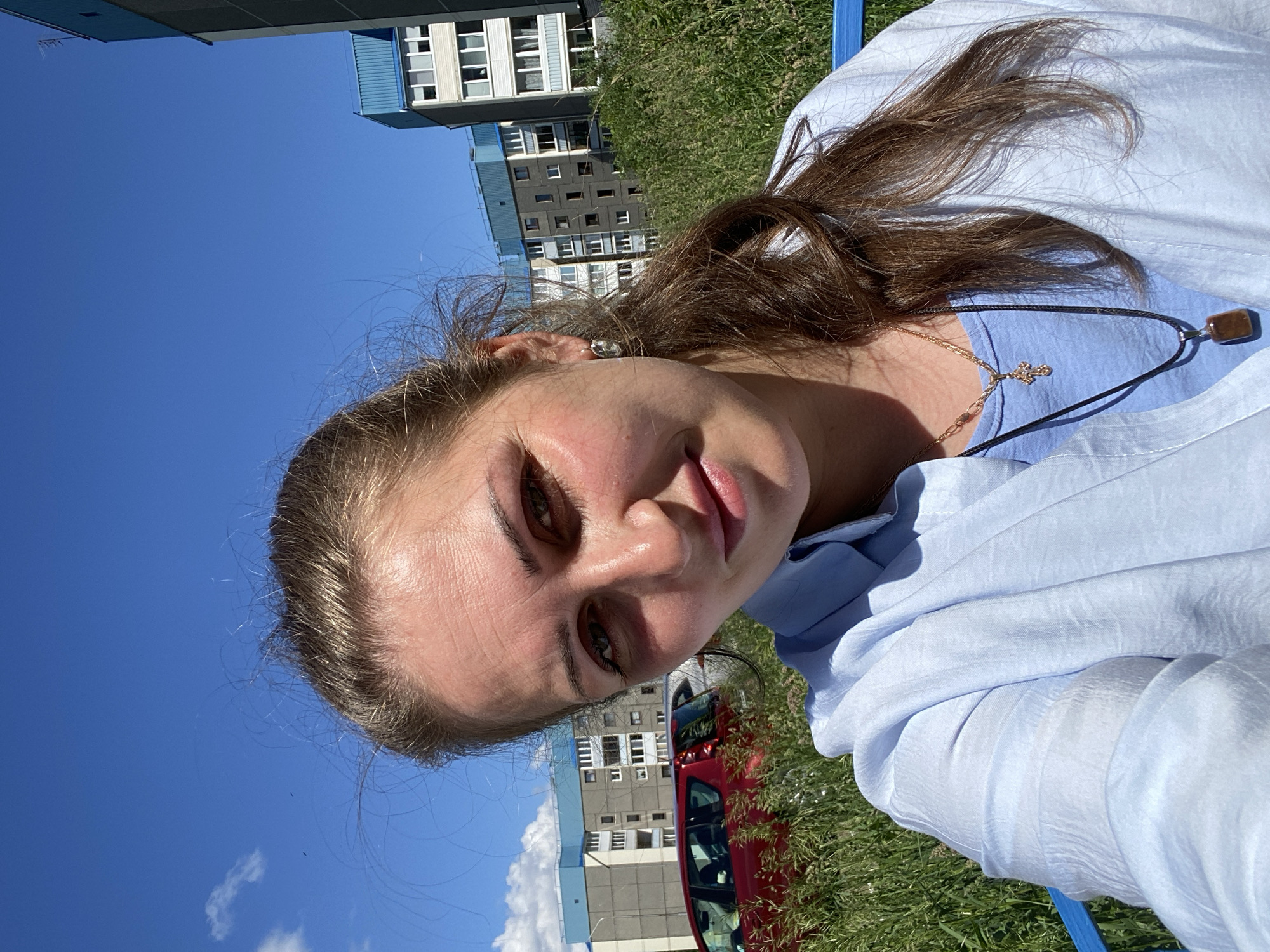 A woman is taking a selfie outdoors on a sunny day. She has light brown hair pulled back in a ponytail and is wearing a light blue collared shirt over a blue t-shirt. She has a cross necklace with a small pendant and another necklace with a rectangular pendant. She is also wearing earrings. The background shows tall grass, a red car, and several apartment buildings with blue accents, consistent with the architecture found in the city of Markovsky, Russia. The sky is clear blue with a few wispy clouds.