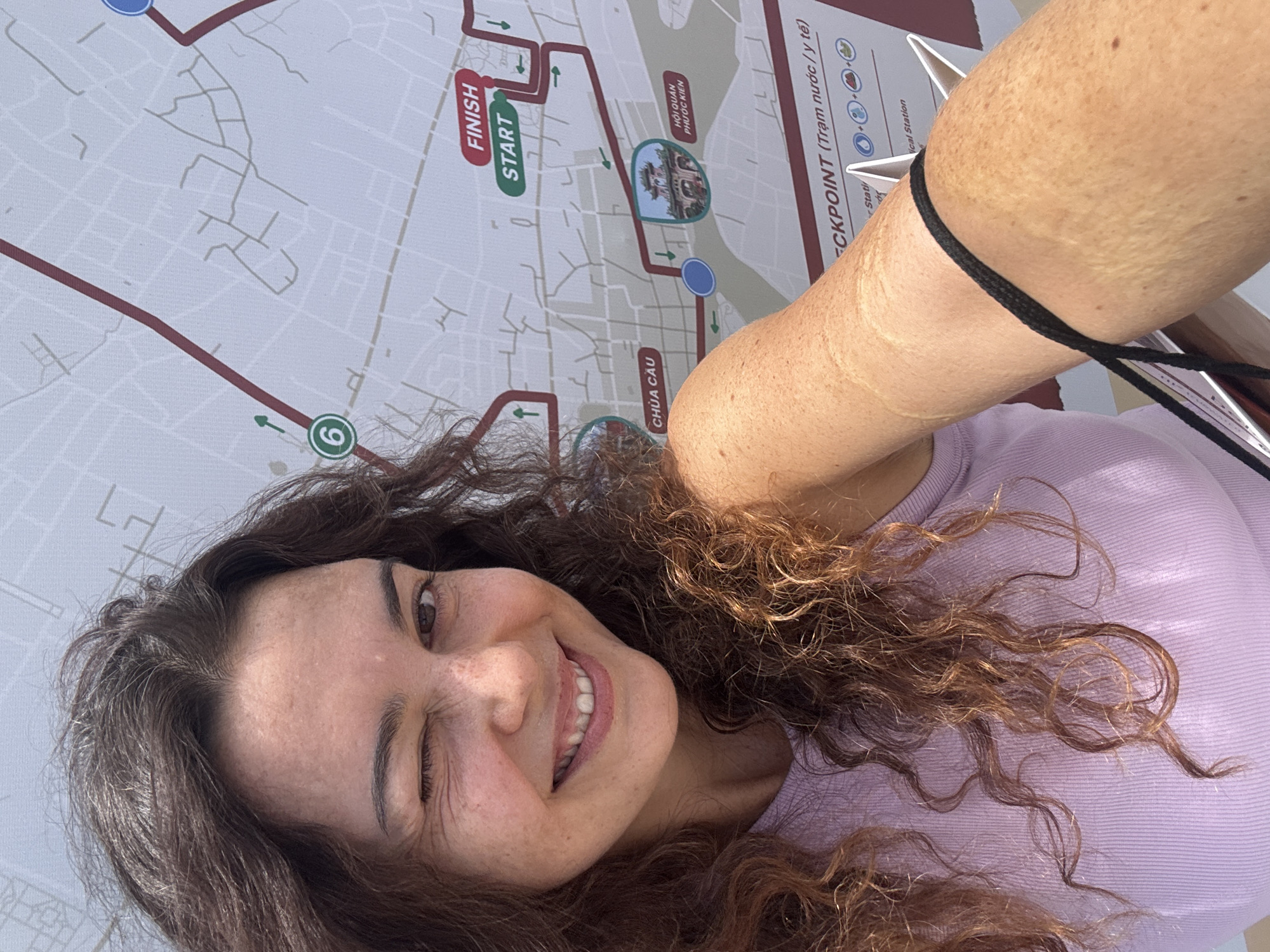 A woman with curly brown hair and freckled skin winks at the camera while standing in front of a map of Hoi An, Vietnam. The map displays a race course with markers for "START," "FINISH," and "CHECKPOINT." She is wearing a purple tank top. The woman's right arm is visible, showing freckles and a black cord tied around her bicep. The map includes Vietnamese text, indicating it is likely a map for a local event or race. The overall lighting suggests an outdoor setting during the day.