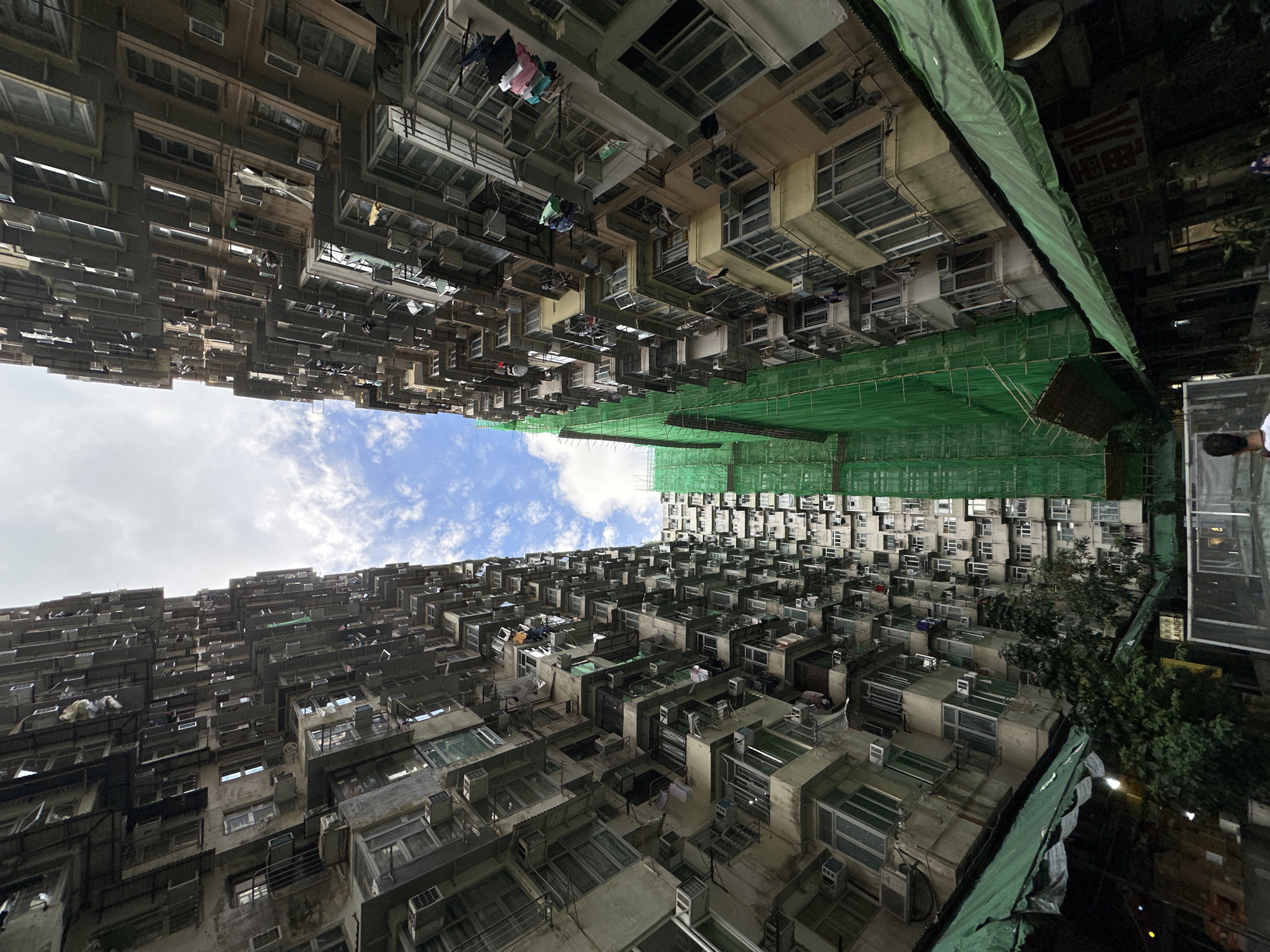 The image depicts a densely populated residential area in Hong Kong, China, characterized by extremely high-rise buildings and a narrow perspective looking upwards towards the sky. Two buildings dominate the composition, their facades crammed with numerous windows and air conditioning units. Clotheslines with laundry are visible between some of the buildings. Construction is occurring, as scaffolding and green protective netting covers parts of the building on the right. A patch of blue sky with scattered clouds is visible between the buildings. Some greenery, possibly trees, is visible on the right side. There appears to be a reflection of a person near the right side.