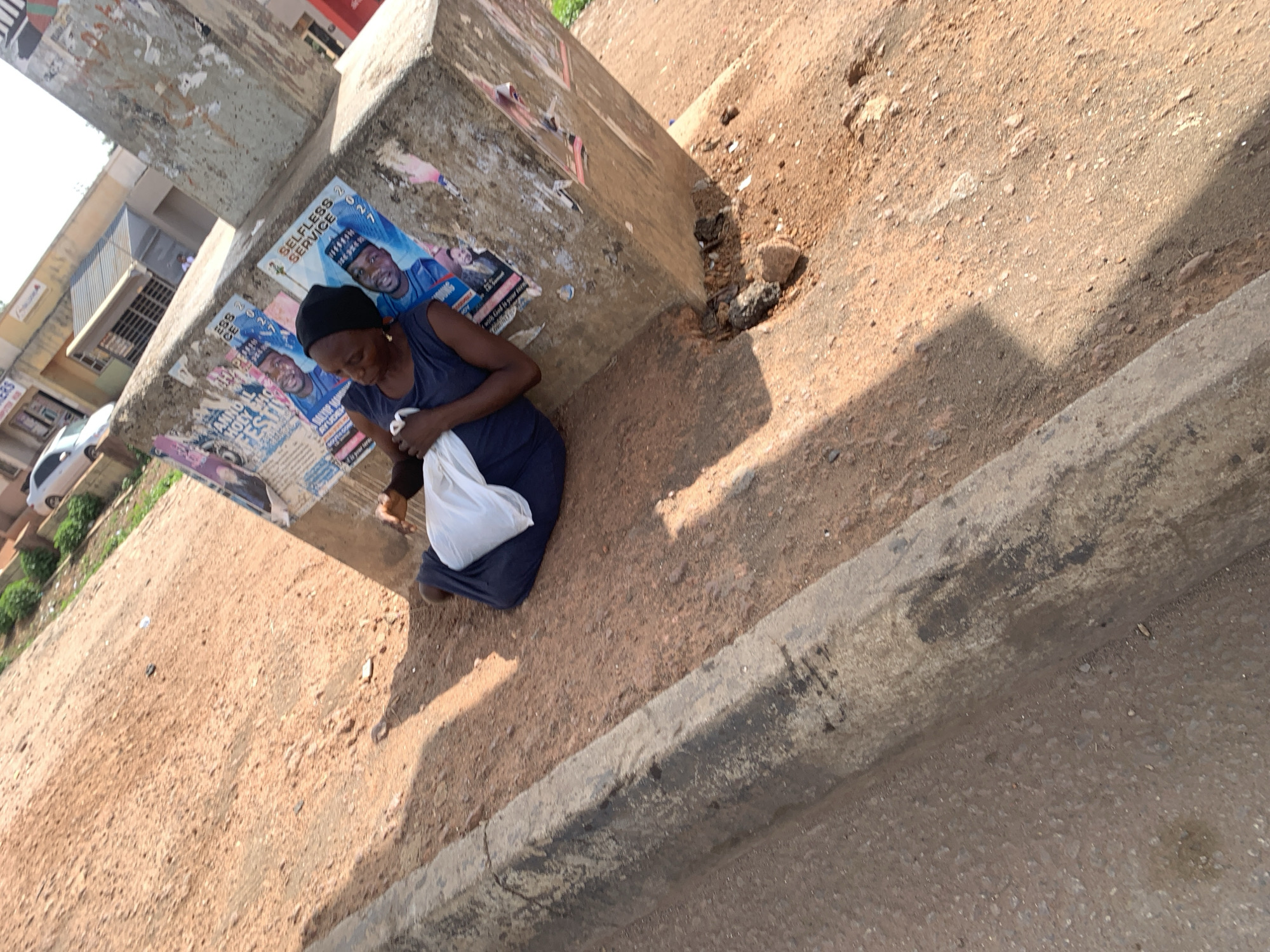 The image shows a woman seated on the ground beside a concrete pillar in Gura Top, Nigeria. She is wearing a dark dress and a head covering and holds a white plastic bag. The pillar is covered in posters, one of which reads "SELFLESS SERVICE." The ground is a mix of dirt and debris, next to a sidewalk or road. Buildings and vehicles are visible in the background.