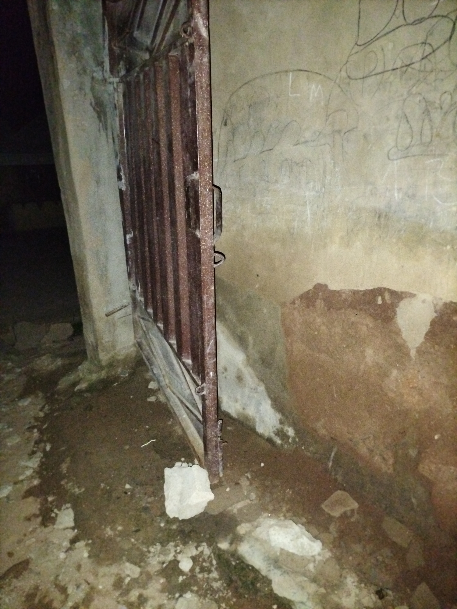 The image depicts a rusted metal gate, partially ajar, leading into a dimly lit area. The gate is made of vertical bars and shows signs of decay. To the left of the gate, a concrete pillar or wall is visible, with some grime and discoloration. The ground in front of the gate is uneven and covered with debris, including rocks and dirt. The background wall, to the right of the gate, is textured and covered in what appears to be graffiti or markings. The lighting is dim, creating shadows and a somewhat unsettling atmosphere. The scene appears to be an outdoor or semi-outdoor location, possibly an alleyway or the entrance to a derelict structure. The location is identified as being in Jalingo, Nigeria.