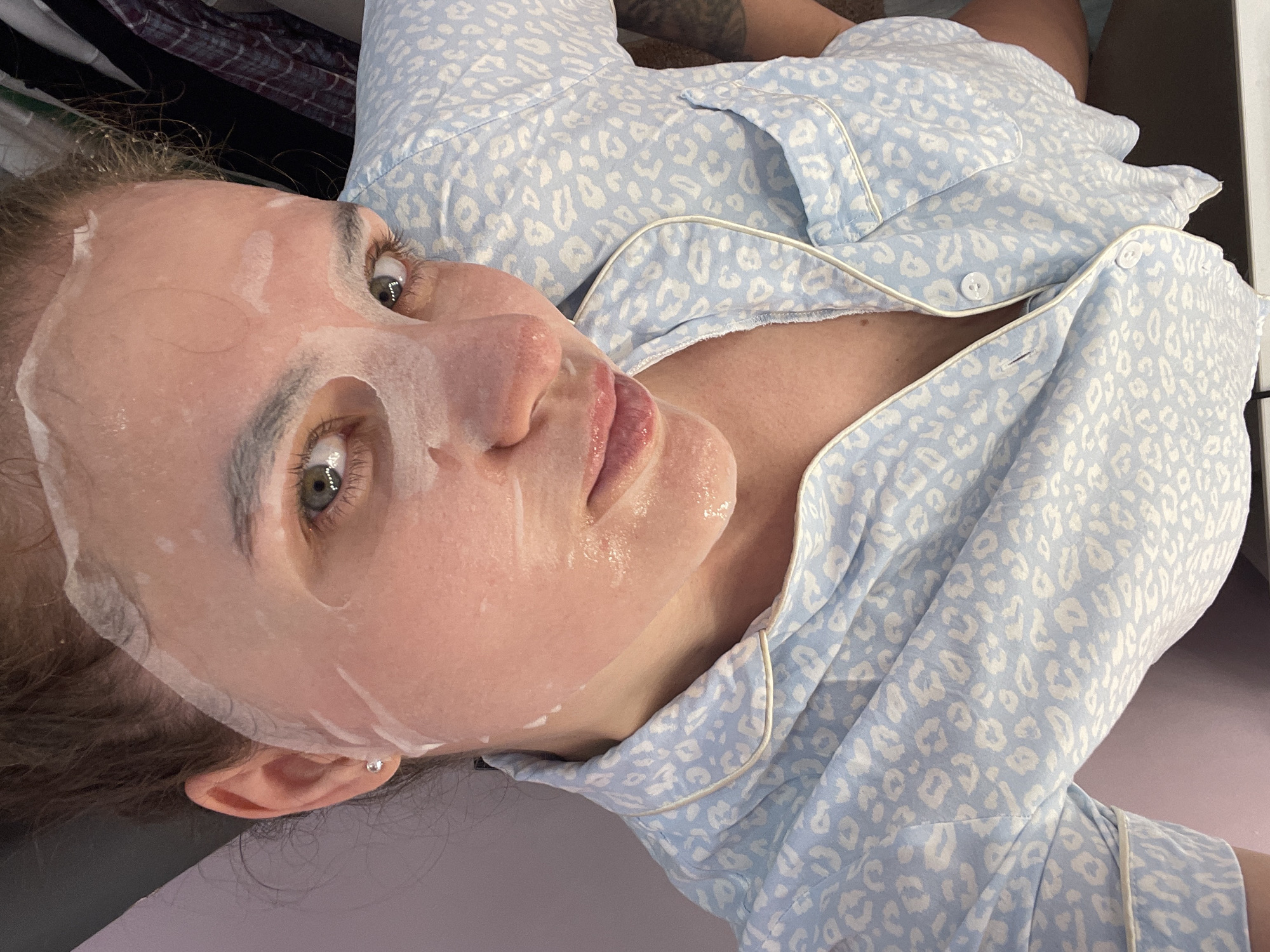A woman with fair skin and green eyes lies on a purple surface, wearing a light blue pajama top with a white leopard print and white piping. Her face is covered by a clear sheet mask. She is looking upwards and towards the left. The location is not discernible from the image.
