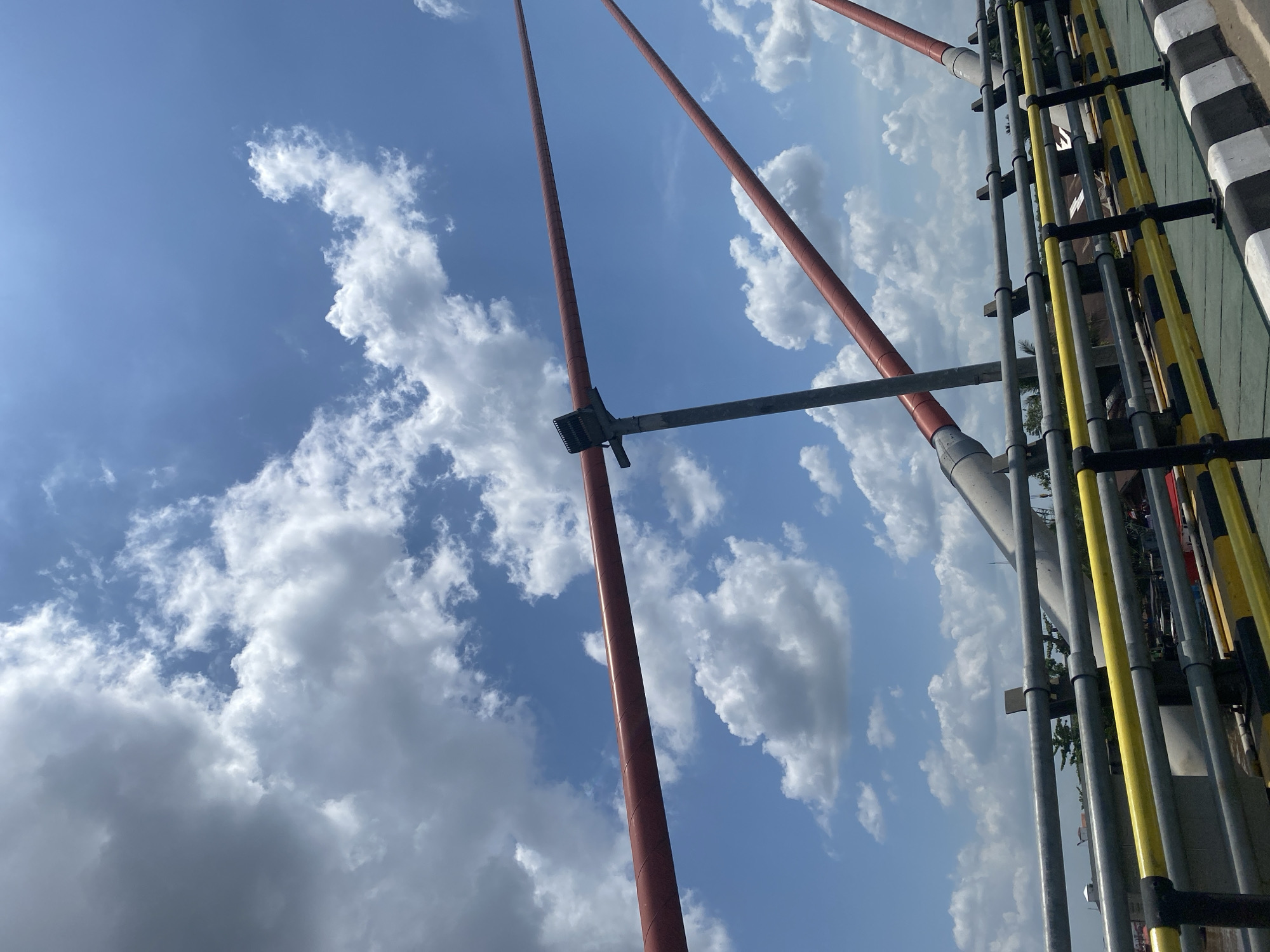 The image shows a section of a bridge with a clear blue sky and white clouds overhead. Red, cylindrical suspension cables are visible, stretching diagonally across the frame, supported by a grey pole with a light fixture. Below the cables, a section of the bridge railing is visible, with horizontal grey bars and yellow and black stripes. The railing is designed to prevent people or vehicles from falling off the bridge. In the background, hints of greenery and buildings can be seen, suggesting an urban environment. The perspective is from within the bridge, looking upwards and outwards.
This bridge is likely the Jembatan Antasan Bromo in Banjarmasin, Indonesia, known for its distinctive red suspension cables and its location over the Martapura River.