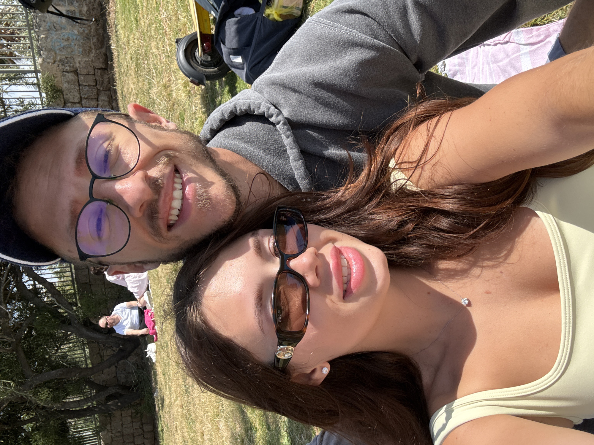 A rotated selfie captures a man and woman lying on grass, likely in a park, potentially Gan Meir in Tel Aviv, given the visible stone wall and trees typical of the area. The man, on the left, wears glasses and a baseball cap, and sports a grey hooded sweatshirt. He is smiling broadly. The woman, on the right, wears sunglasses and a light-colored top. A silver necklace with a pendant is visible on her neck. An electric scooter is parked nearby, partially obscured by a bag, suggesting personal transportation. Other people are in the background, engaged in leisure activities.