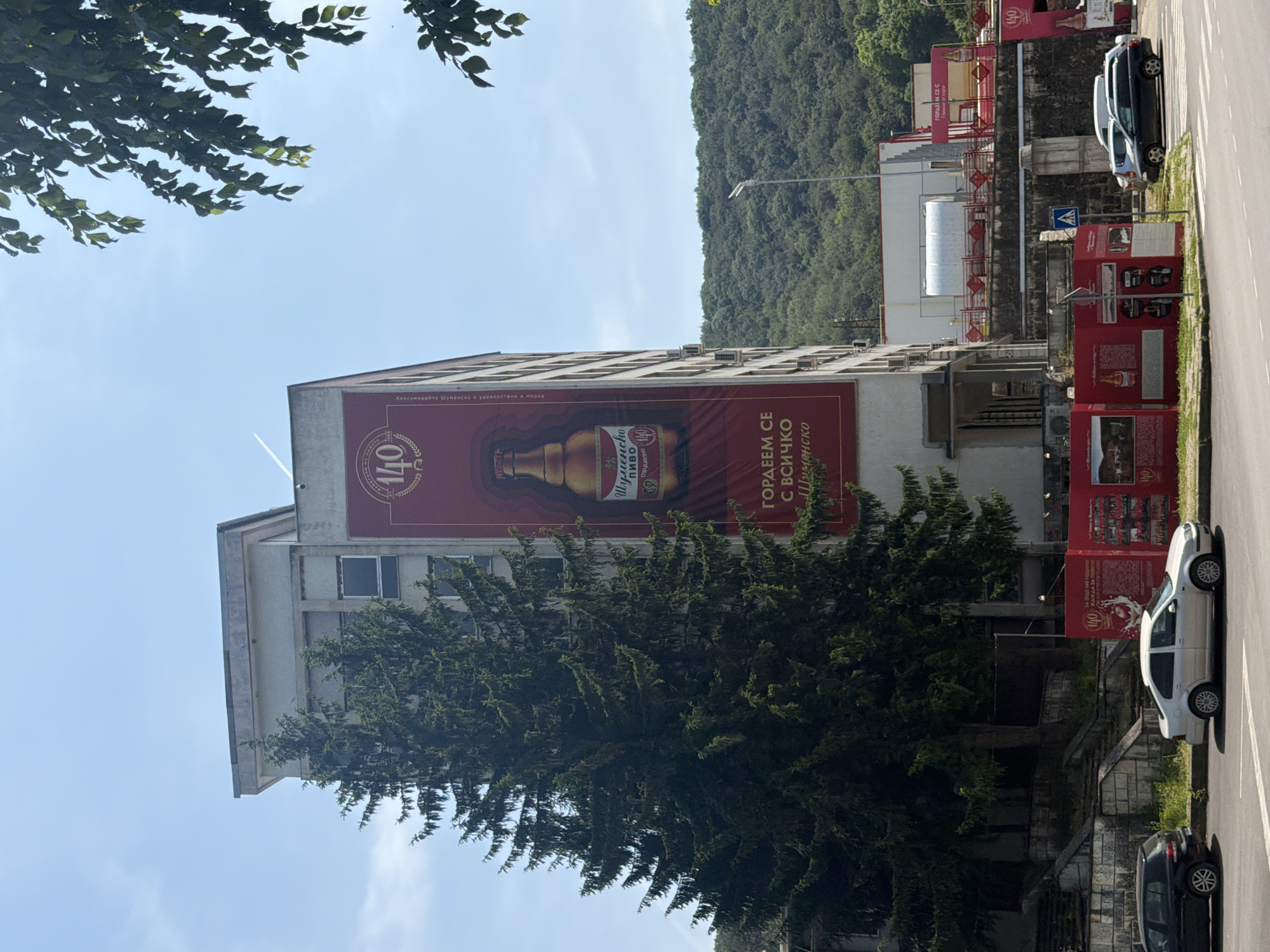The image depicts a multi-story, light-colored building under a clear blue sky, located in Shumen, Bulgaria, during bright daylight. The building's right facade is largely covered by a prominent red banner celebrating "140 ГОДИНИ" (140 Years) of "ШУМЕНСКО ПИВО" (Shumen Beer), featuring a large golden beer bottle and the slogan "ГОРДЕЕМ СЕ С ВСИЧКО ШУМЕНСКО" (We are proud of all things Shumen).
To the left of the building stands a tall, dense evergreen tree, obscuring much of the building's lower and mid-section on that side. Air conditioning units are visible on the building's facade beneath the banner.
In the foreground, a paved road with white lane markings runs horizontally. A dark grey hatchback and a silver sedan are parked on the left side of the road, facing right. Further right, a black station wagon and a white van are parked. Along the sidewalk, red metal barrier fencing is erected, featuring several informational panels adorned with the "140" logo, text in Bulgarian, and historical images, indicating a temporary exhibition or construction barrier related to the brewery's anniversary. A pedestrian crossing sign is visible behind these barriers.
Behind the parked cars on the left, a set of concrete stairs and a retaining wall lead up to an elevated area. In the mid-ground, beyond the road and structures, a modern streetlamp is visible. The background consists of densely forested green hills under the bright sky. A few green tree branches extend into the upper right corner of the frame. A faint white contrail is visible in the sky above the building. No individuals are discernible in the scene.