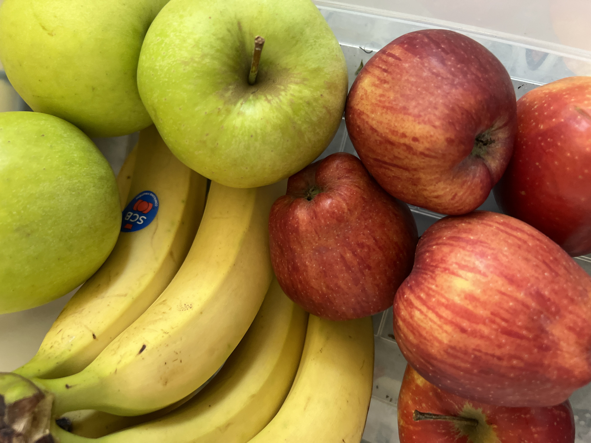 A top-down, close-up shot features a collection of fresh fruit, including bananas and apples. The bananas are bright yellow with a few brown spots and are arranged in a bunch at the top left of the frame. Some of the bananas have blue and white stickers on them. To the right of the bananas are several green apples, their surfaces smooth and taut. Below the bananas and interspersed with the green apples are several red apples, their skins mottled with red and yellow hues, some showing faint stripes. The fruit appears to be placed on a white surface, possibly a countertop or a tray.