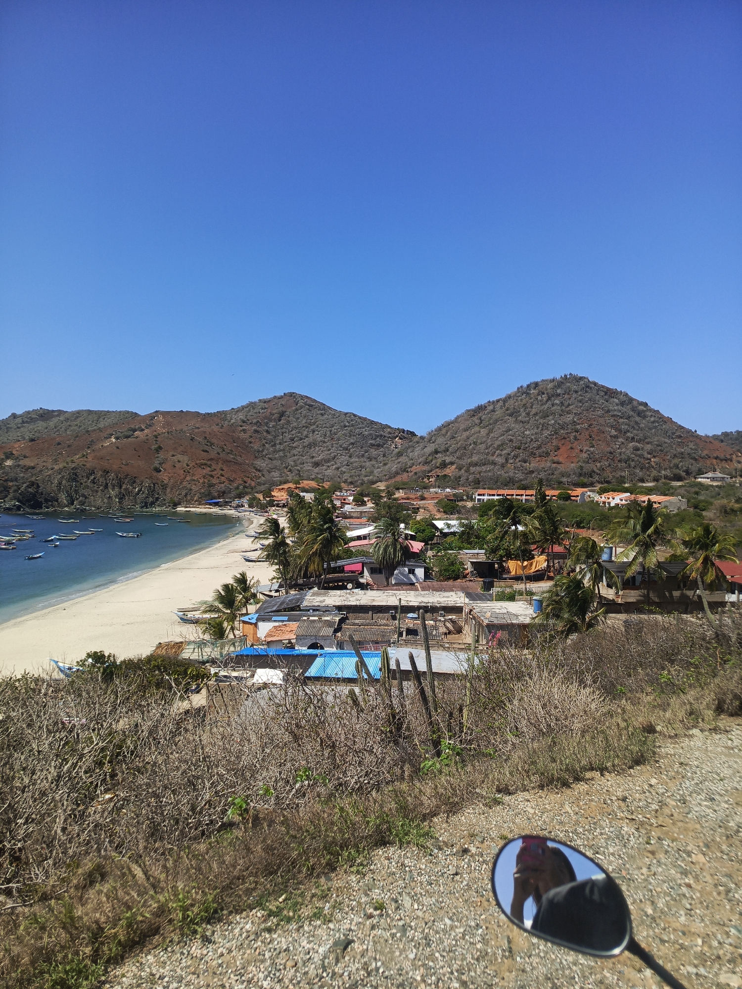 A high-angle, medium shot captures a coastal village nestled between arid hills and a calm, blue body of water. The beach is a stretch of light sand, populated with numerous small, dark boats anchored offshore. Along the coastline, a cluster of buildings with red and blue rooftops forms a small settlement. Palm trees are scattered throughout the village. The landscape is predominantly dry and rocky, with sparse vegetation covering the hills. The sky is a clear, bright blue. In the foreground, a motorcycle mirror reflects the image of a person wearing a pink headscarf and a dark jacket. The scene appears to be in the city of Manzanillo, Venezuela, based on its characteristic geography and architecture.