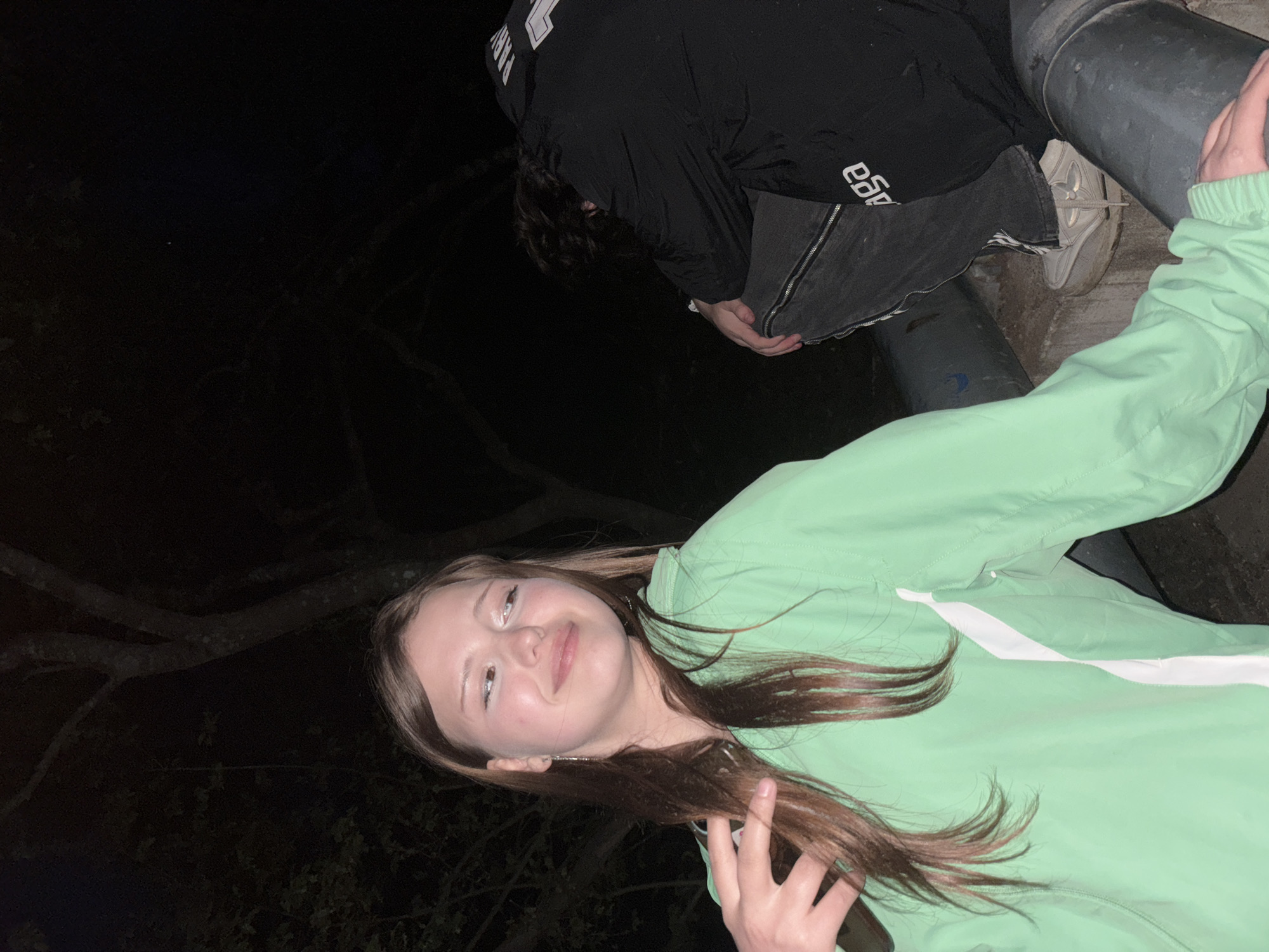 The photograph, taken at night in an outdoor location in Фоки, Russia, features two individuals. A young woman is positioned closer to the foreground, wearing a light green jacket and facing the camera with a slight smile. Her hand is partially visible in the bottom right corner of the frame. Behind and above her, a second individual is present, bending over or seated on what appears to be a concrete or stone structure. This person is dressed in a dark jacket with white lettering, paired with grey pants and white sneakers. The background is largely dark, with branches of trees visible on the left side of the image. There are also grey pipes/tubes/structures behind them. The location appears to be an urban or semi-urban area based on the presence of man-made structures.