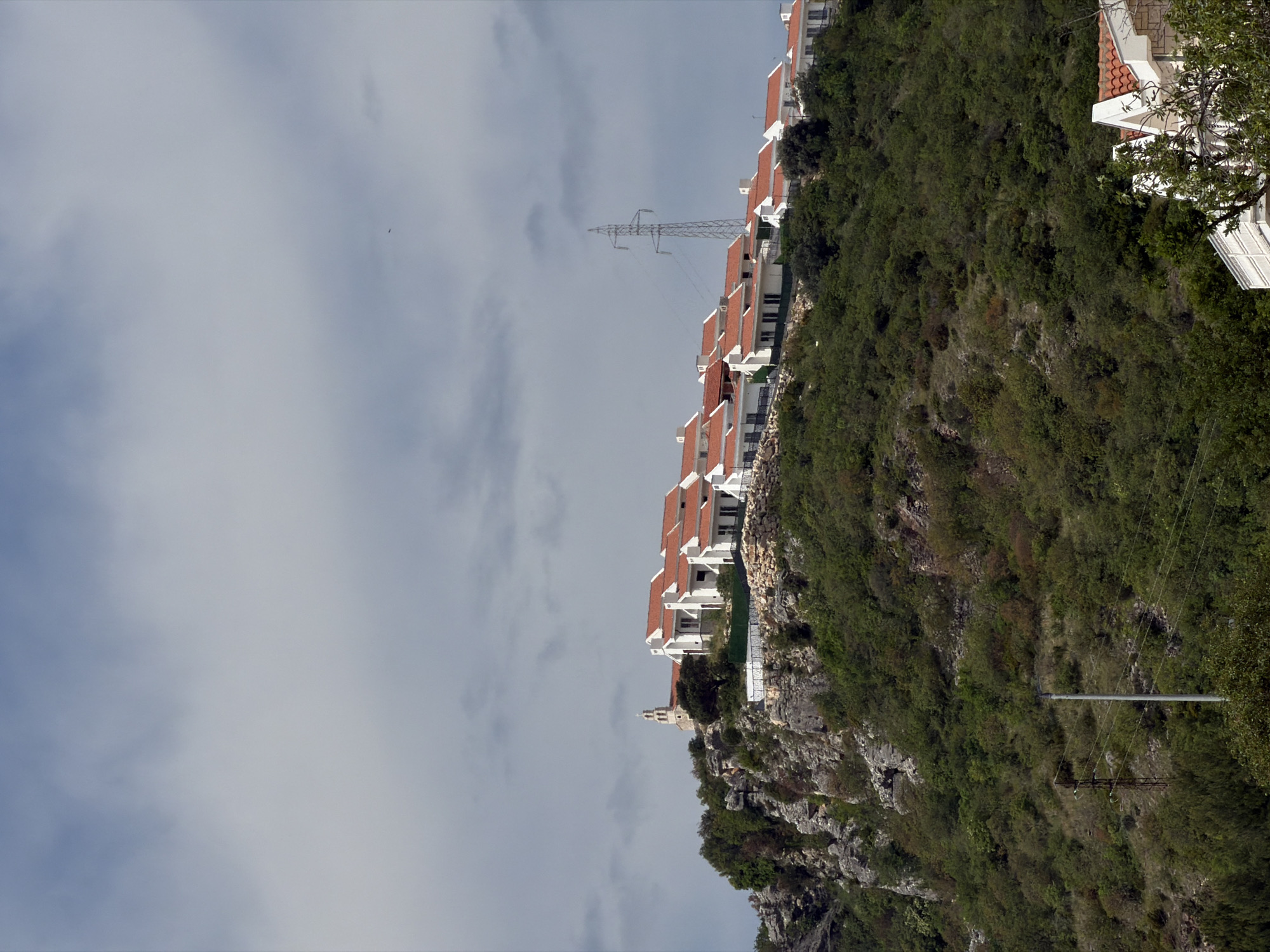 The image depicts a hillside in Blizikuće, Montenegro, with a row of modern white villas with red-tiled roofs perched along the top edge of the slope. Below the villas, the hillside is covered with dense green vegetation and rocky outcrops. In the distance, an electrical transmission tower stands against a cloudy sky. At the bottom of the hillside, a church steeple can be seen amongst the trees. Power lines are visible along the hill.