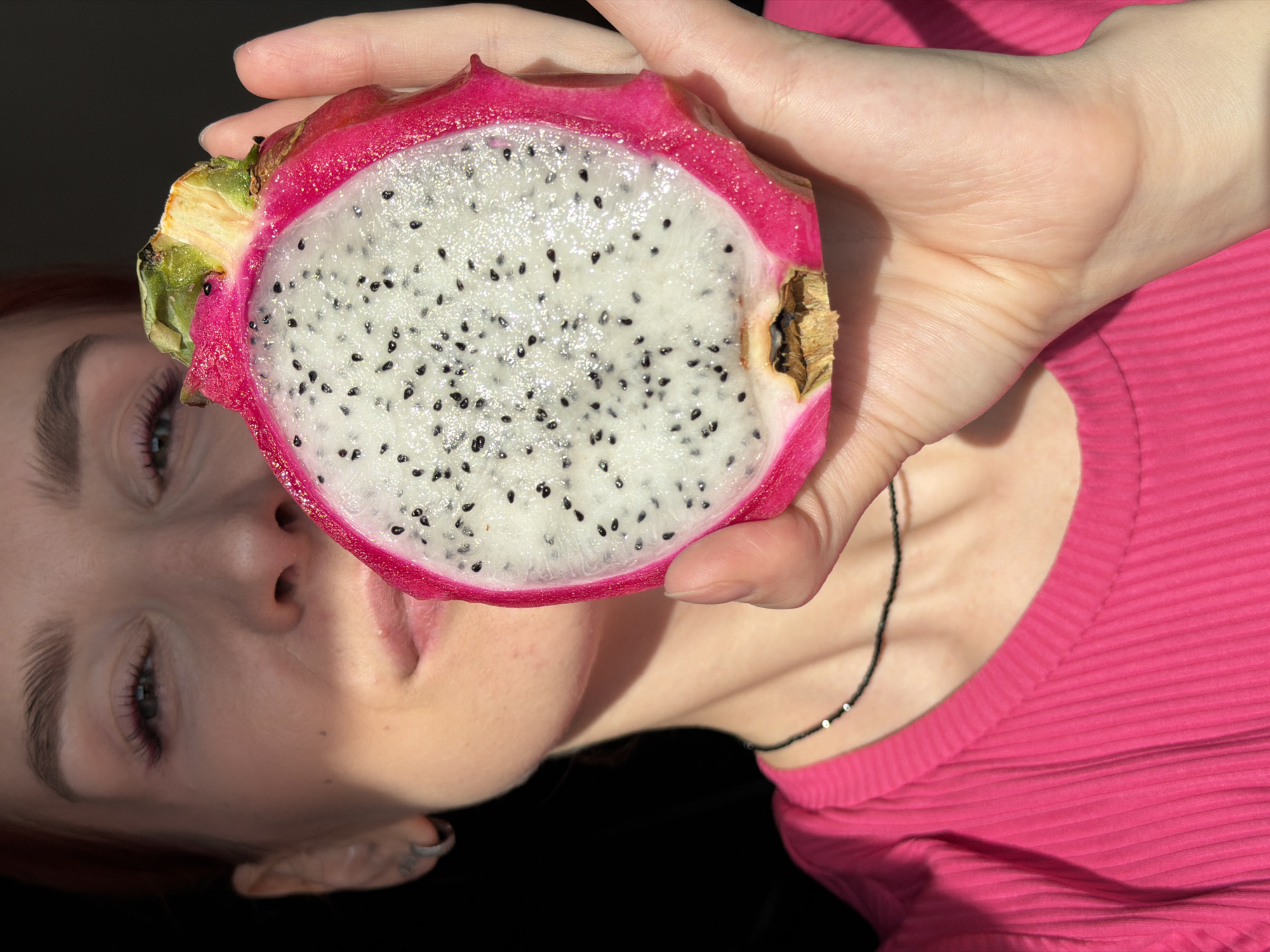 A fair-skinned woman with reddish hair is holding a halved dragon fruit up to her face. The fruit is cut in half, revealing its white flesh speckled with tiny black seeds. The outer skin of the fruit is pink with green leaves on the top. The woman is wearing a hot pink ribbed shirt and a black beaded necklace. The background is dark and blurry. The image appears to be taken indoors with artificial lighting. There is no visible indication of the location being Chaikovsky, Russia.