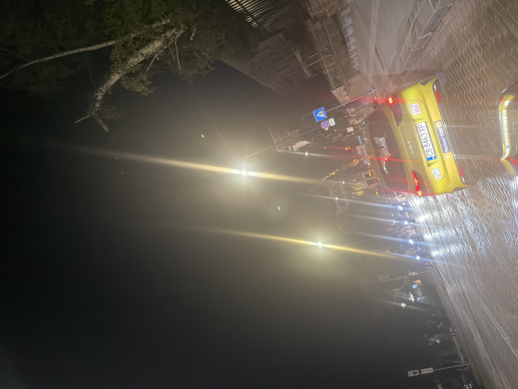 The image depicts a night scene in Sofia, Bulgaria, featuring a yellow Kia car with the license plate CB 8463 XP parked on a street with a cobblestone-like surface. The car has a "Yellow! App" advertisement on it, suggesting it is a taxi. There are other vehicles and traffic lights visible in the background. Road signs are also present. A tree branch overhangs the scene from the top left of the frame. A building or structure appears above the cars. The overall lighting is low, with significant glare and lens flare from artificial light sources.