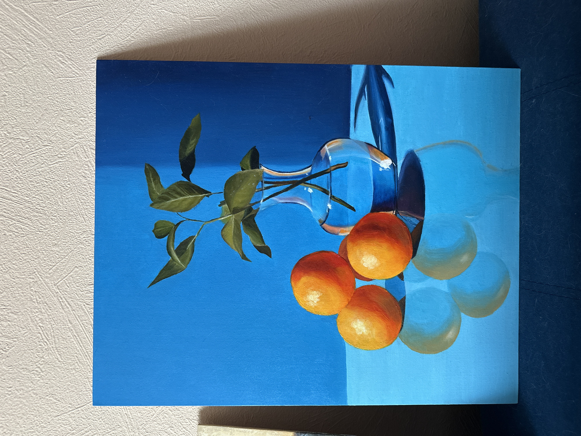 A still life painting depicts oranges, a glass vase, and green foliage against a divided blue background. The painting is photographed indoors, leaning against a wall. The location is not discernible from the image.