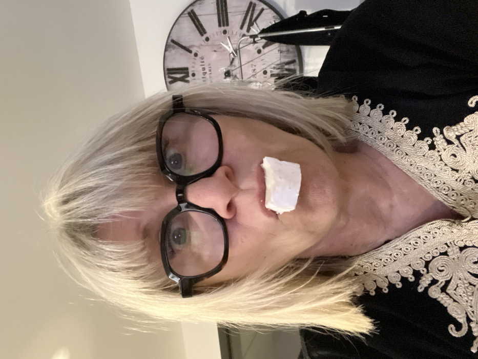 A woman with short, light blonde hair and black-framed glasses has a small, white, possibly square piece of cheese stuck to her upper lip. She's wearing a black top with ornate cream-colored trim around the neckline. A decorative clock is visible in the background, slightly out of focus. The image appears to be taken at a tilted angle.