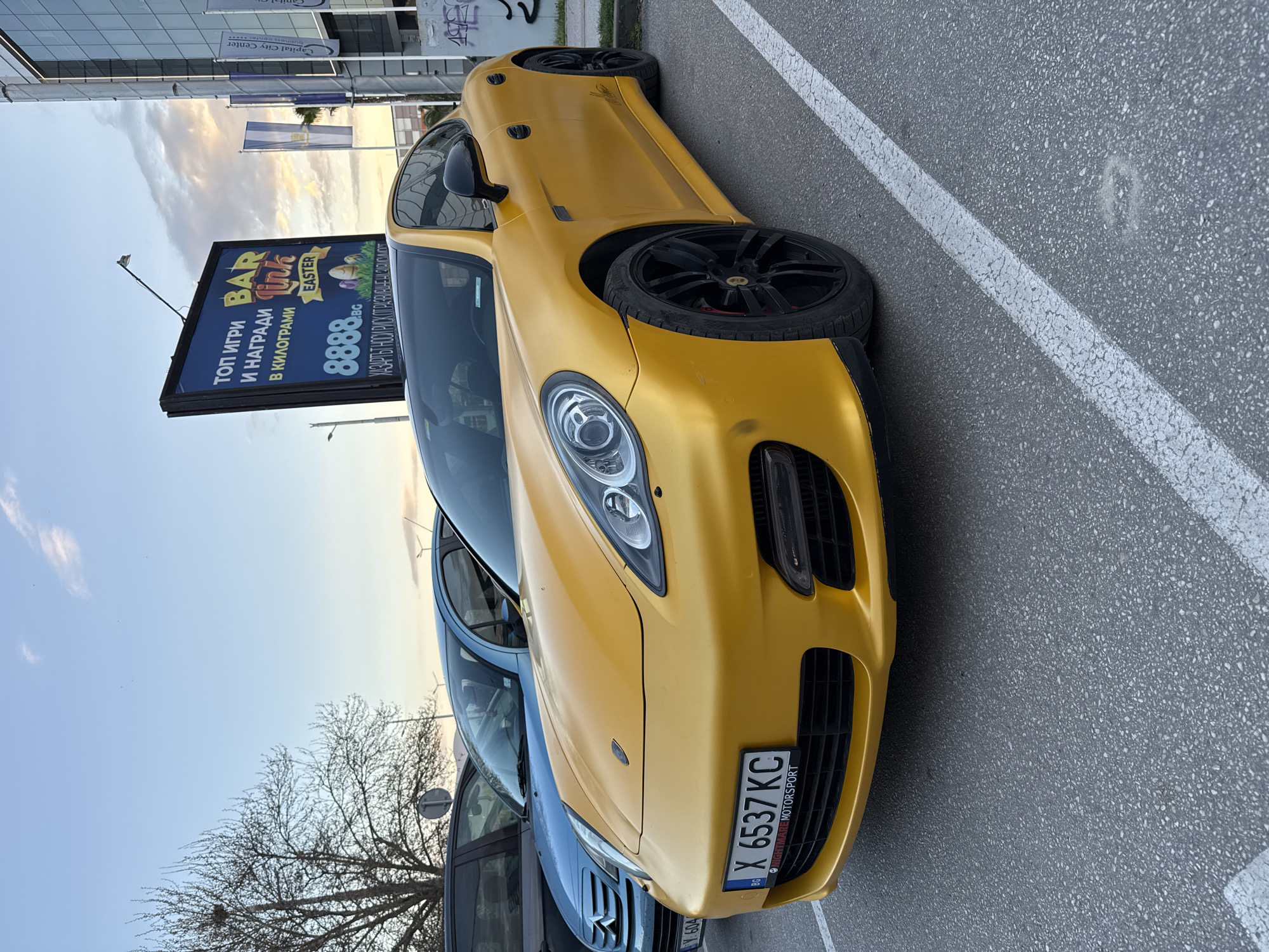 The image depicts a matte gold Porsche parked in a parking space adjacent to a white painted line on asphalt. The car has black rims and a license plate reading "X 6537 KC" with the words "NIGHTMARE MOTORSPORT" beneath. Next to the Porsche is a blue car partially visible in the shot. In the background is a billboard advertising "BAR Link EASTER" and "ТОП ИГРИ И НАГРАДИ В КИЛОГРАМИ" with the number "8888.BG." Above the billboard, a building with the text "Capital City Center" can be seen. The sky is visible with shades of blue and orange suggesting either sunrise or sunset. Bare tree branches are in the lower left. It is impossible to verify whether this image was shot in the city of Plovdiv, Bulgaria.