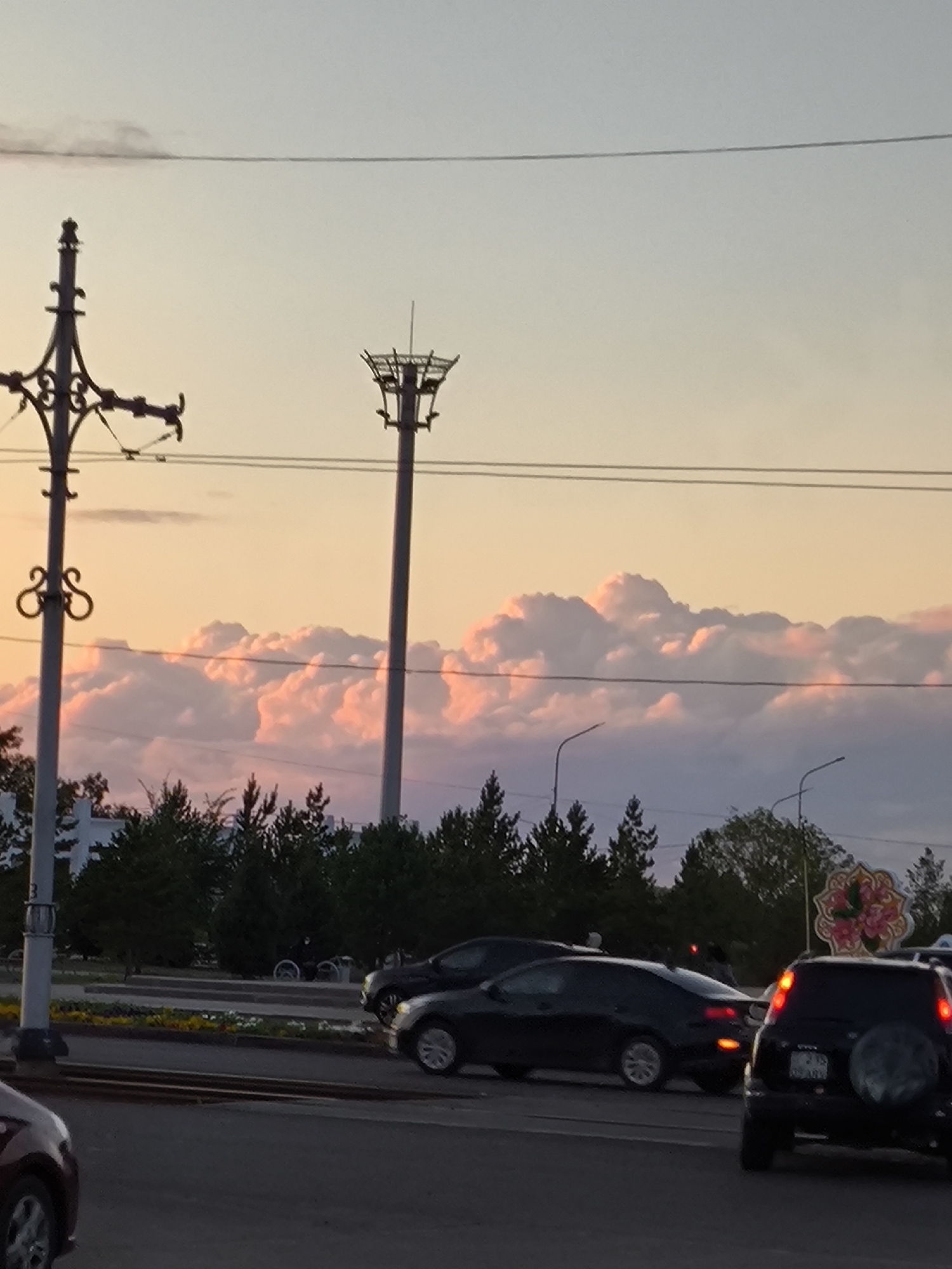 This is a medium-angle, eye-level shot of a street scene in Temirtau, Kazakhstan, during a sunset. The sky is filled with large, pink and white cumulus clouds, illuminated by the setting sun. A tall, industrial-looking tower with a platform at the top stands prominently in the mid-ground, to the right of a decorative lamppost on the left. Utility wires stretch across the sky. In the foreground, vehicles are present on a multi-lane road. A black SUV is visible on the right, with its brake lights illuminated. To its left, a black coupe is driving, its taillights also lit. A portion of a maroon car is visible in the bottom left corner. Trees line the background, with some buildings and streetlights also discernible. The overall impression is that of an urban environment at dusk, characterized by industrial infrastructure and natural beauty.