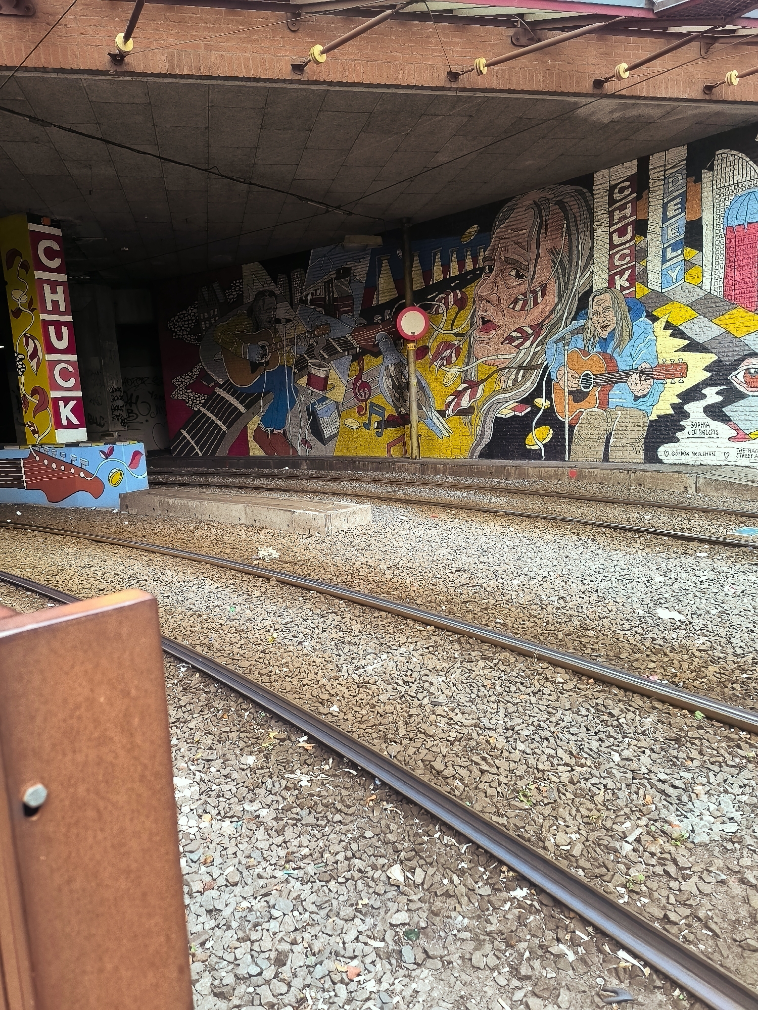 This is a photograph taken at the KKN-tunnels under the Buitenhof in The Hague, Netherlands. The image captures a section of a vibrant mural adorning the tunnel walls, featuring a depiction of musicians playing guitars, with stylized faces and architectural elements. To the left, a vertical "CHUCK" text artwork is visible on a yellow pillar. Train tracks run through the foreground of the tunnel, indicating it's a functional transit area. The ground is covered with gravel and debris. The overall scene is an urban environment characterized by street art and infrastructure.