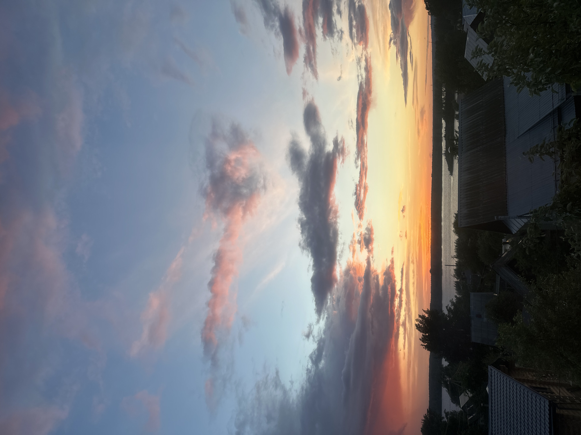 The image shows a sunset over a body of water. The sky is filled with clouds, with hues of orange, pink, and blue. The water reflects the colors of the sky. On the right side of the image, there are buildings and trees silhouetted against the sunset. It is not possible to confirm the location within Chaikovsky, Russia, or any specific details about people, subjects, objects, actions, or interactions within the scene based on this image alone.