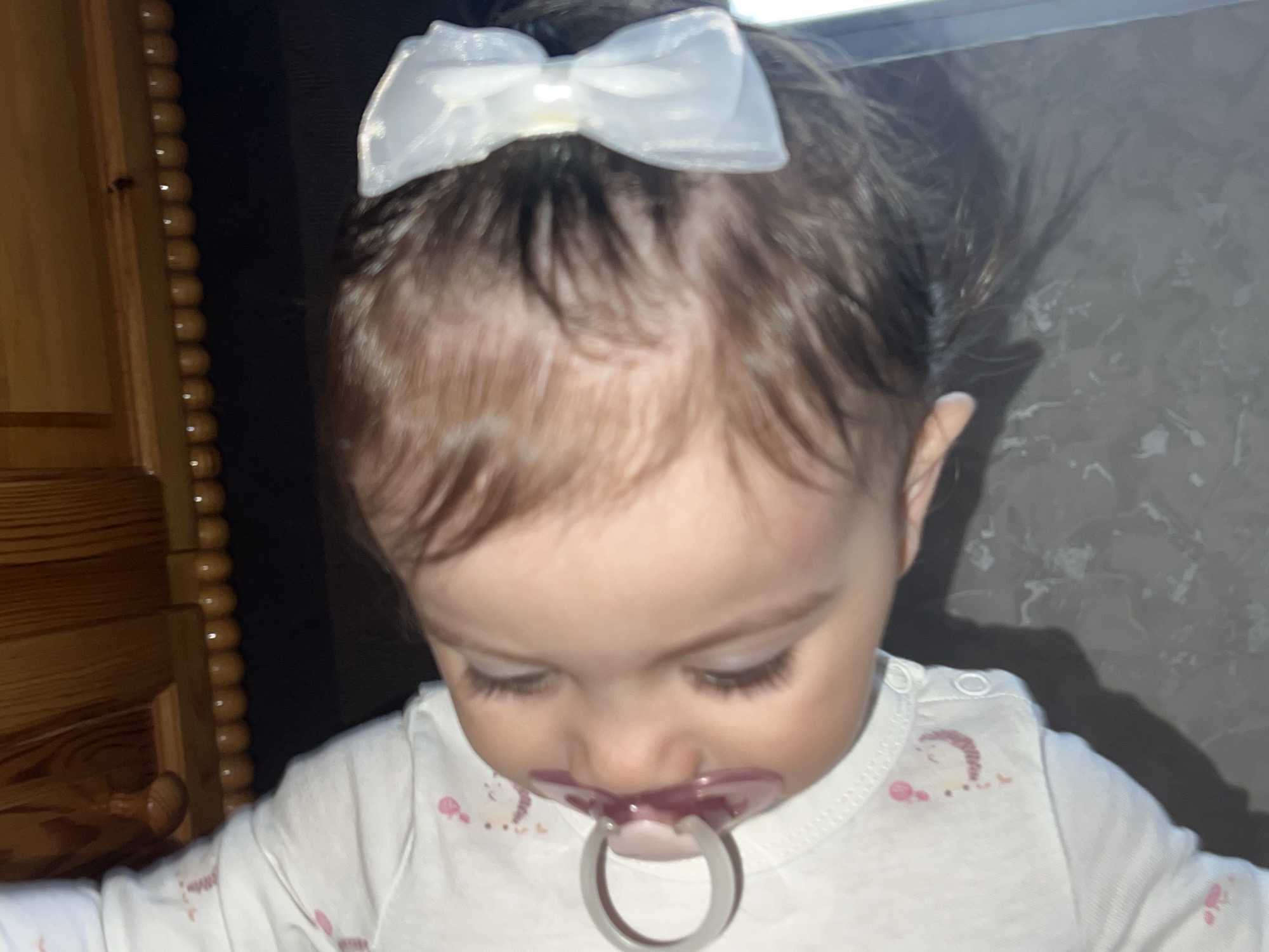 A close-up, high-angle shot captures a baby, likely a girl, indoors in Elena, Bulgaria. The baby is positioned centrally, looking downwards with a neutral, focused expression. She has dark brown hair, which is slightly disheveled, and wears a translucent white bow on top of her head. A purple-tinted pacifier is in her mouth. Her long eyelashes are clearly visible as she gazes down.
She is dressed in a white long-sleeved onesie or top, featuring faint light pink designs, including a rainbow-like arc on her right shoulder and abstract splotches or shapes across the fabric. White snap buttons are visible on the shoulder of her garment.
To the left of the frame, a section of light brown wooden furniture or decor is visible, characterized by a vertical line of rounded, bead-like embellishments. The background to the right of the baby is predominantly dark, appearing to be a wall or dark surface, with some subtle variations in tone and texture. The lighting is soft and appears to be artificial, highlighting the baby's features and accessories. No discernible text is visible in the image.