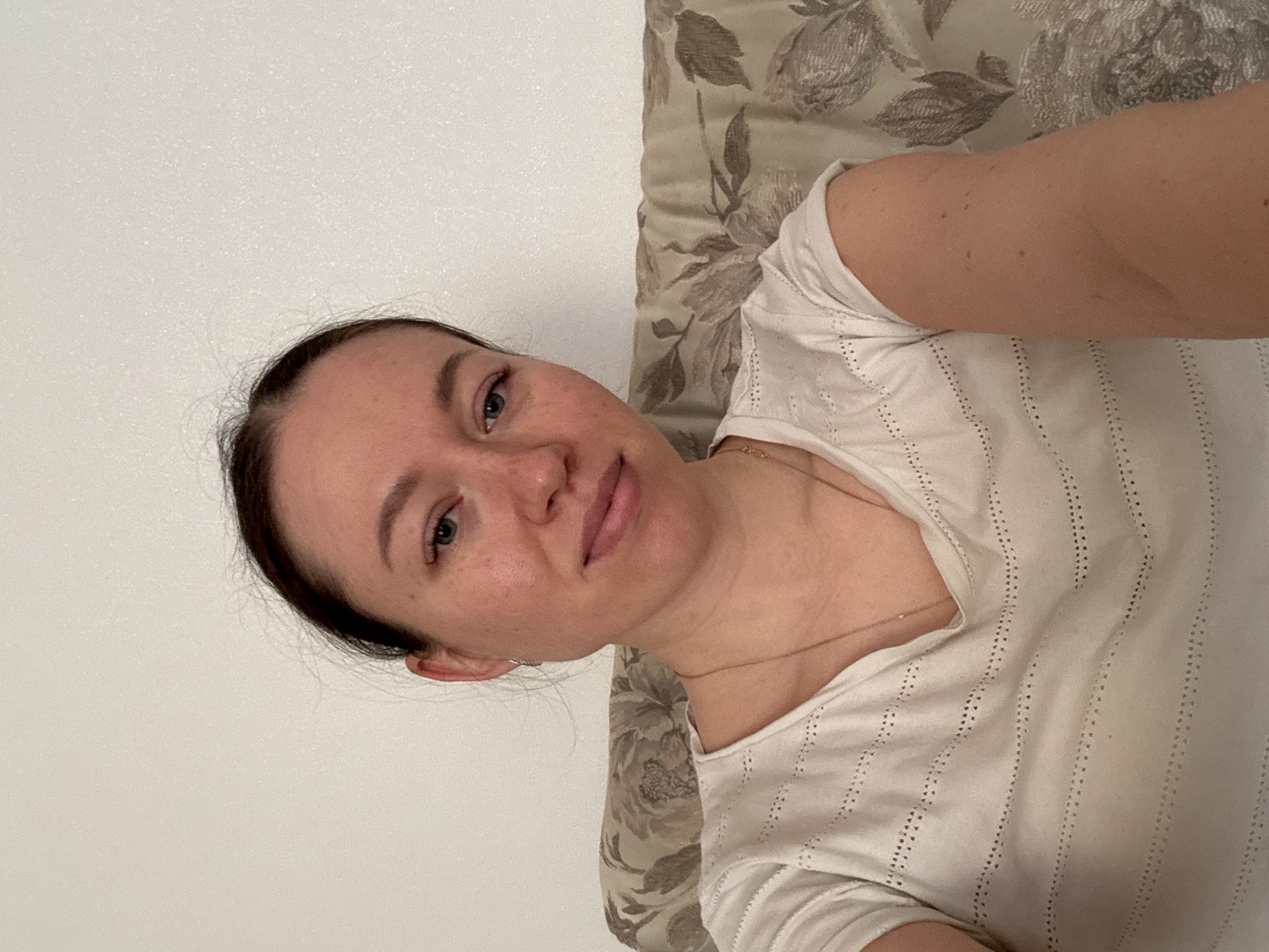 A woman with dark hair and light skin is lying down on a floral patterned cushion or pillow, positioned next to a white wall. She is wearing a white shirt with small vertical lines of cutouts. A thin gold necklace is visible around her neck. The image appears to be a selfie. There is no information available to confirm that the image was taken in Ферма, Russia.