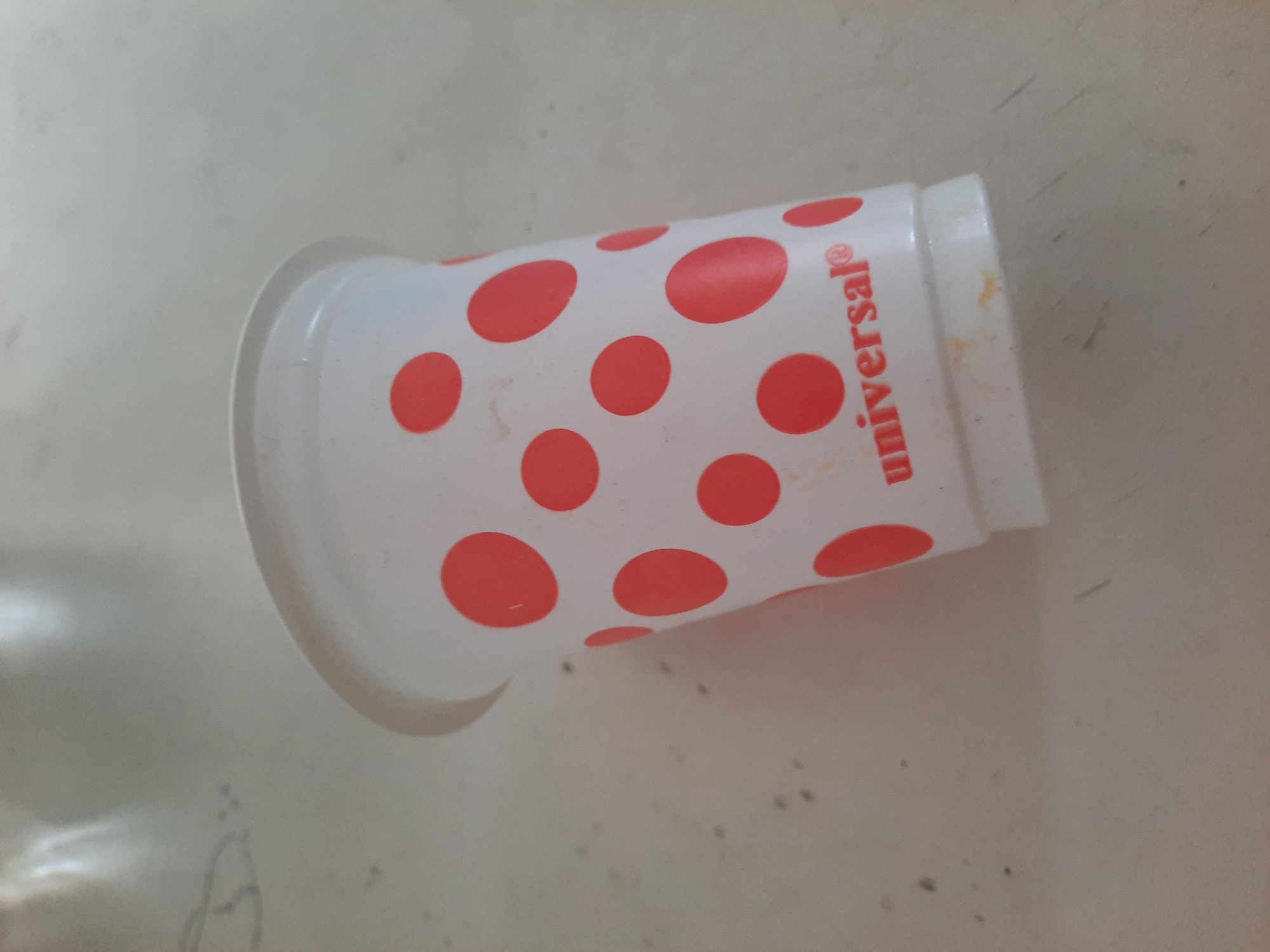 The image shows a white plastic cup with red polka dots, branded with the word "universal®" in red lettering. The cup appears to be disposable and has a lid. It is resting on a light-colored, textured surface, possibly a table, which has some smudges and debris. The lighting suggests an indoor setting. There is no indication of the cup's location in Lhokseumawe, Indonesia.