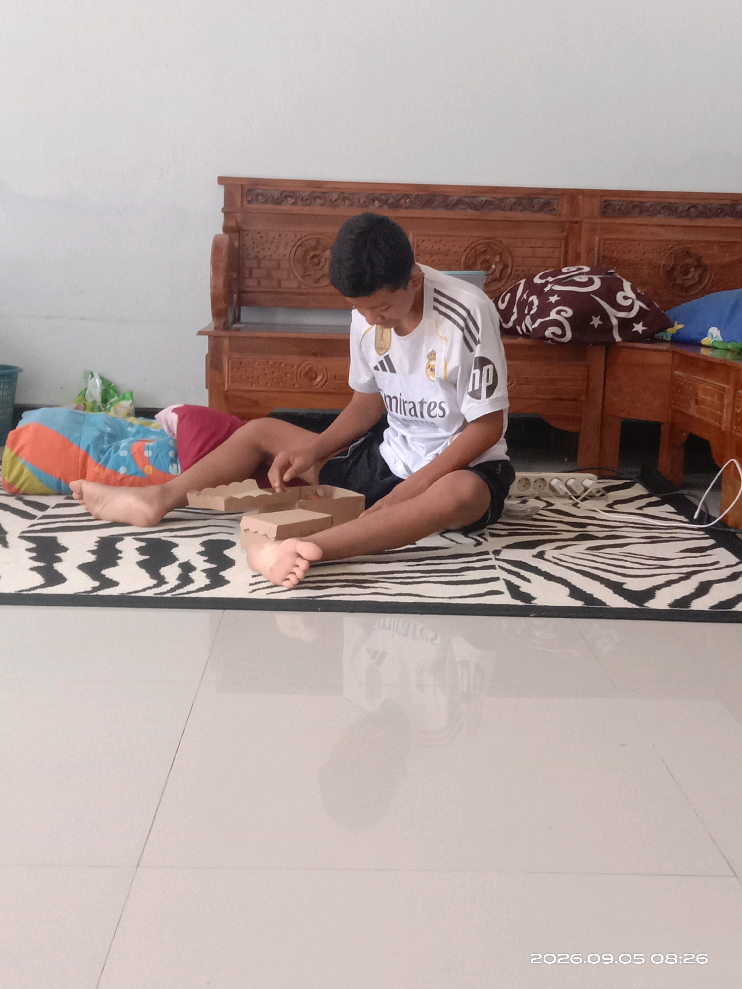 The image depicts a young male sitting on a zebra-patterned mat on a tiled floor, indoors. He is wearing a white jersey with "Emirates" printed on it and black shorts. The boy is engaged in an activity with two small brown cardboard boxes in front of him. To his left, on the mat, are colorful pillows. In the background, a carved wooden bench is visible, along with a white wall. The overall scene suggests a casual domestic setting. The date stamp at the bottom right of the image indicates "2026.09.05 08:26." The location of Banyuarang, Indonesia is not directly evident from the image itself, but the scene is consistent with an indoor environment in that region.