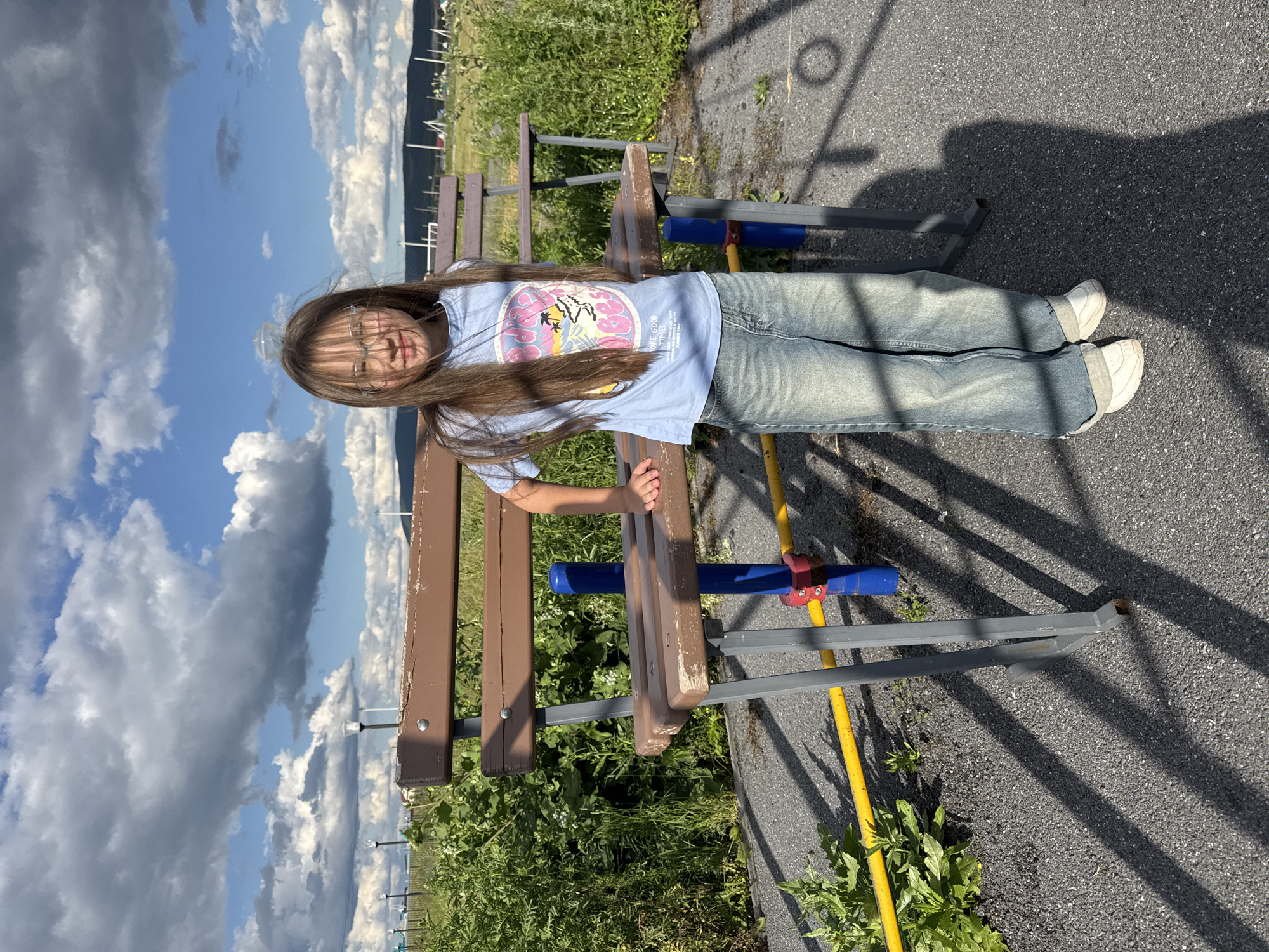A young girl with long brown hair and glasses stands in front of a brown park bench outdoors. She is wearing a light blue t-shirt with a graphic print and loose-fitting blue jeans. The bench has a weathered appearance with visible scratches on the wood. Behind the bench, dense green foliage and taller grasses are visible. The ground in the foreground is paved with dark grey asphalt, marked with the shadow of the bench and what appears to be a fence. A yellow pipe runs horizontally in front of the bench, and a blue vertical pole with red accents is situated beneath it. In the background, a distant landscape features rolling hills, scattered structures, and power lines under a cloudy blue sky. The scene is illuminated by natural daylight.
