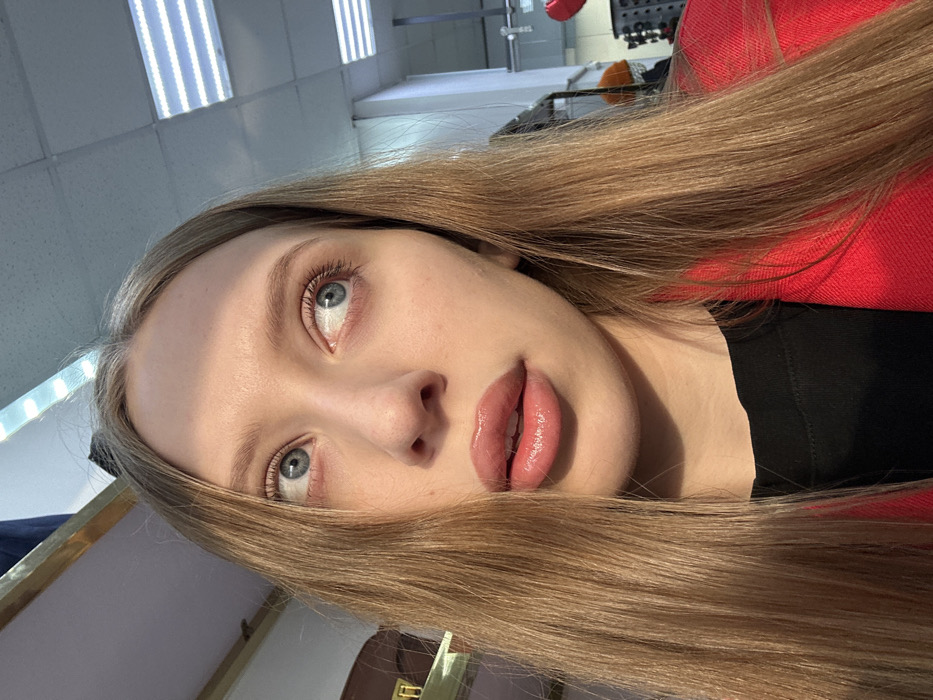 A close-up image captures a female individual indoors in Chaikovsky, Russia. The subject has long, straight, light brown hair and blue eyes, which are directed upwards and to the viewer's left. Her lips are slightly parted, exhibiting a glossy, light pink application. She wears a black top beneath a red blazer. Strong, warm directional lighting illuminates the left side of her face and hair, creating a soft shadow on the right. The background features a white suspended ceiling with multiple rectangular LED light panels. To the left, a partial view of a gold-edged fixture is visible. To the right, obscured elements consistent with a retail store environment are present, including dark display racks and a glimpse of an orange object.