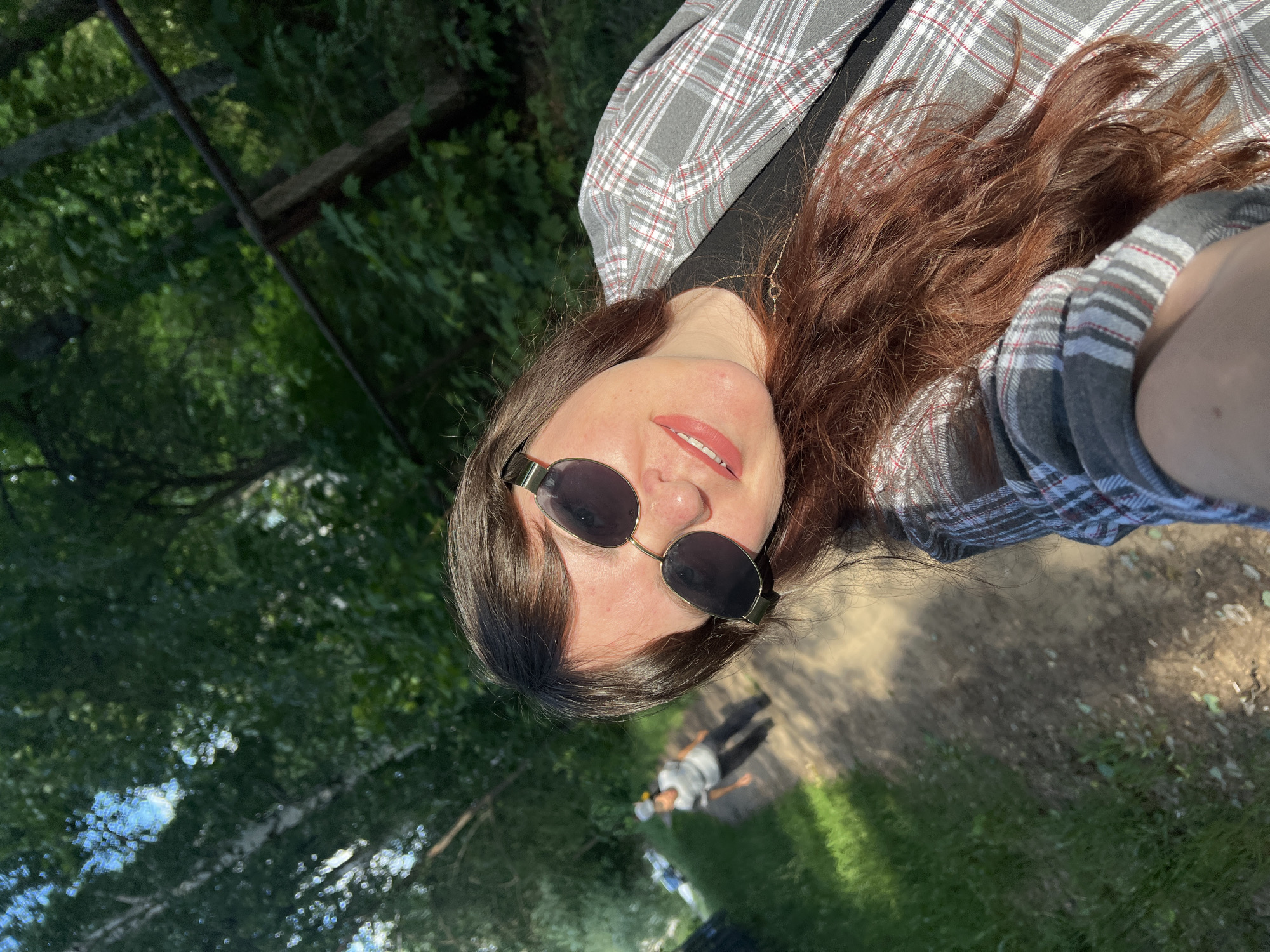 An outdoor, waist-up, selfie style photograph depicts a woman with long, dark brown hair and sunglasses, wearing a grey and white plaid jacket over a black shirt, and a delicate gold necklace. She is smiling and leaning towards the camera. Behind her, a dirt path winds through a wooded area with lush green trees. Sunlight filters through the leaves, casting dappled shadows on the path. In the background, a person wearing a grey shirt and a white hat is walking away from the camera on the path. A partially visible wooden pole with a horizontal bar runs diagonally across the upper right portion of the frame. The scene suggests a natural, possibly recreational, environment.