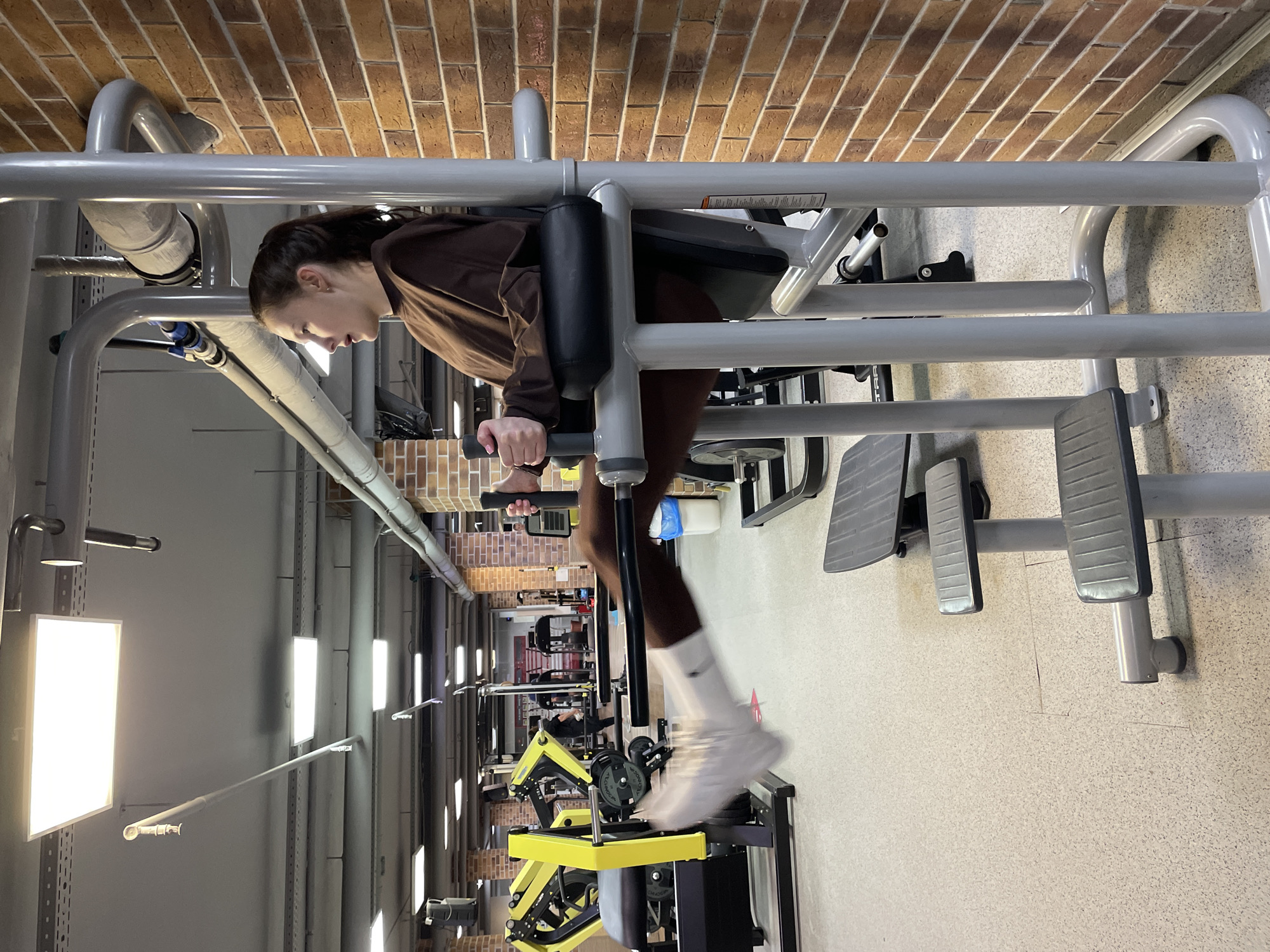 A woman is exercising on a leg press machine in a gym. The gym is located in Izhevsk, Russia. The walls are brick, and the floor is tiled. There are other exercise machines in the background. The woman is wearing a brown sweatshirt and leggings. She is wearing white socks and sneakers. She is holding onto the handles of the leg press machine and pushing the footplates with her feet.